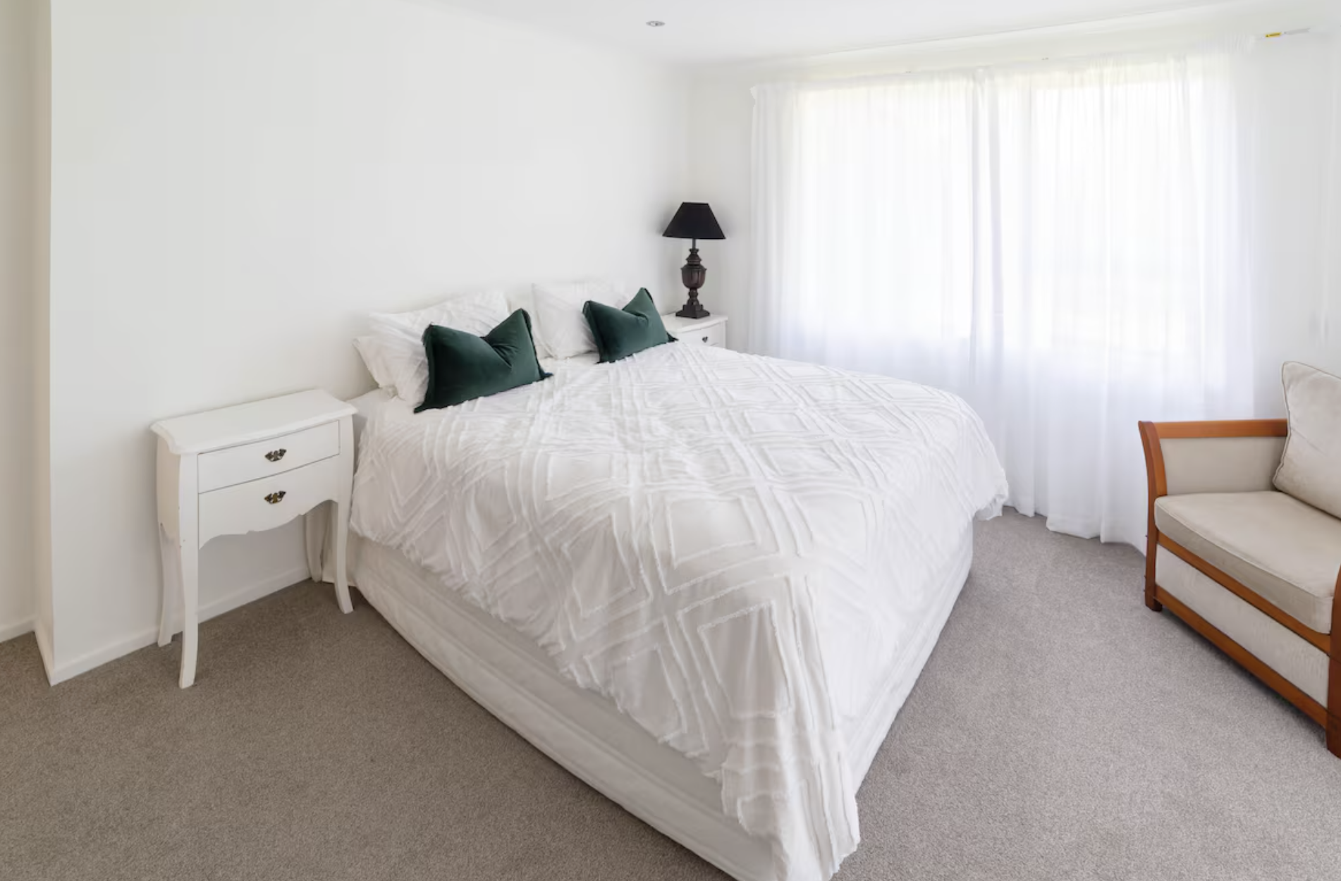 A bedroom with a bed, nightstand, lamp, couch, and large window with white curtains