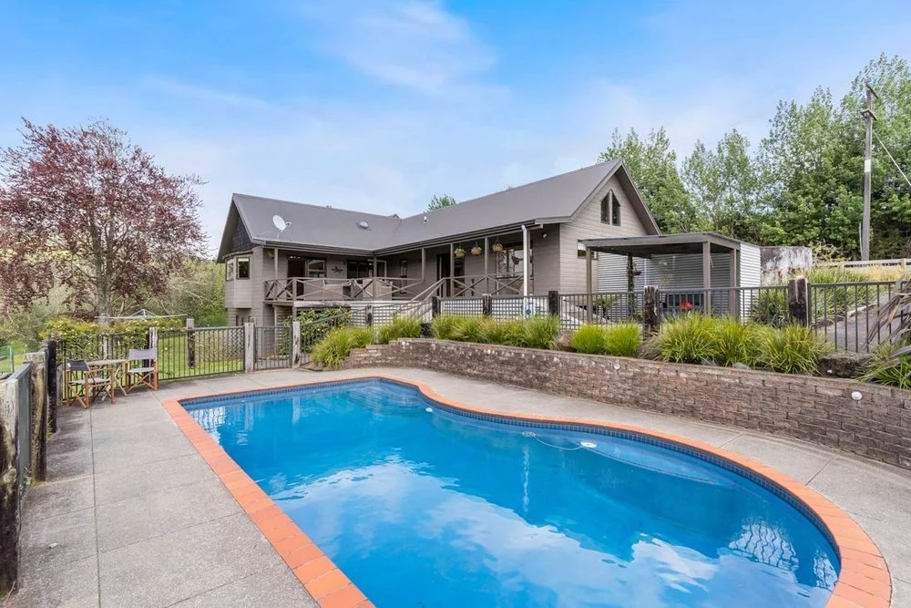 Residential backyard with a swimming pool, patio chairs, a wooden house with a porch, and surrounding greenery.