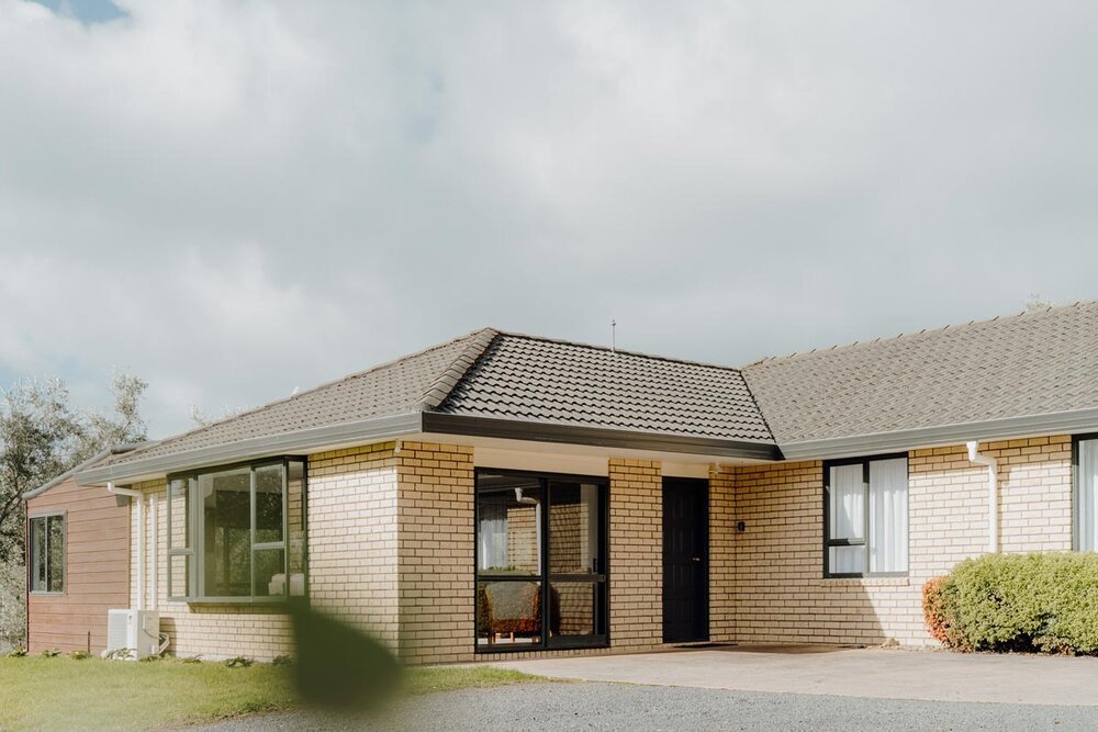 A single-story brick house with a tiled roof, black front door, and large windows, situated on a grassy lawn with shrubs and an overcast sky.