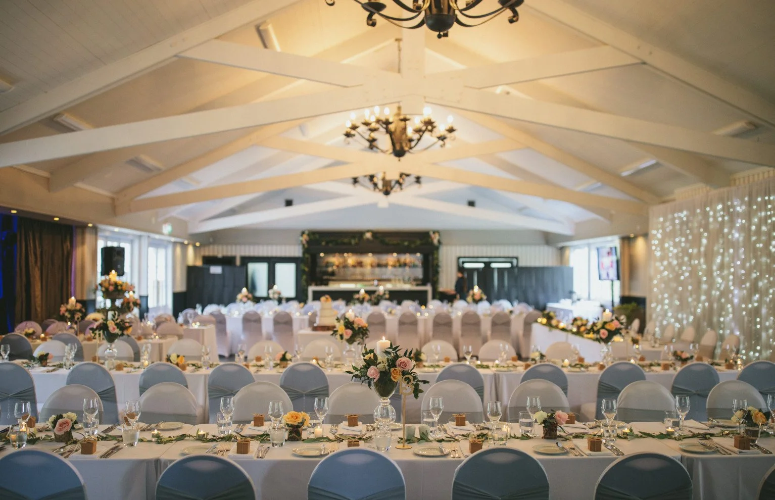 Elegant banquet hall decorated for a wedding reception with long tables set with floral centerpieces, candles, and place settings, and decorated with string lights and flower arrangements.