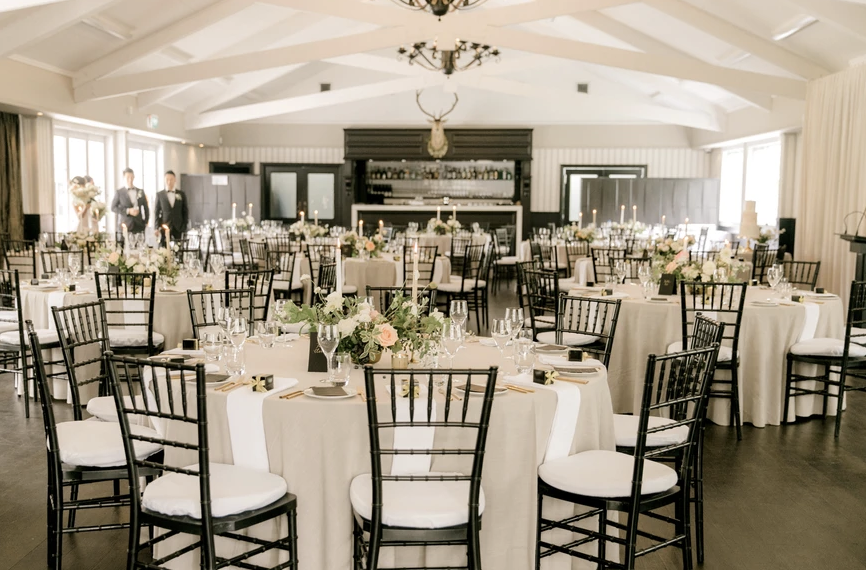 Elegant banquet hall decorated for a wedding reception with round tables, floral centerpieces, black chairs, and a mounted deer head chandelier