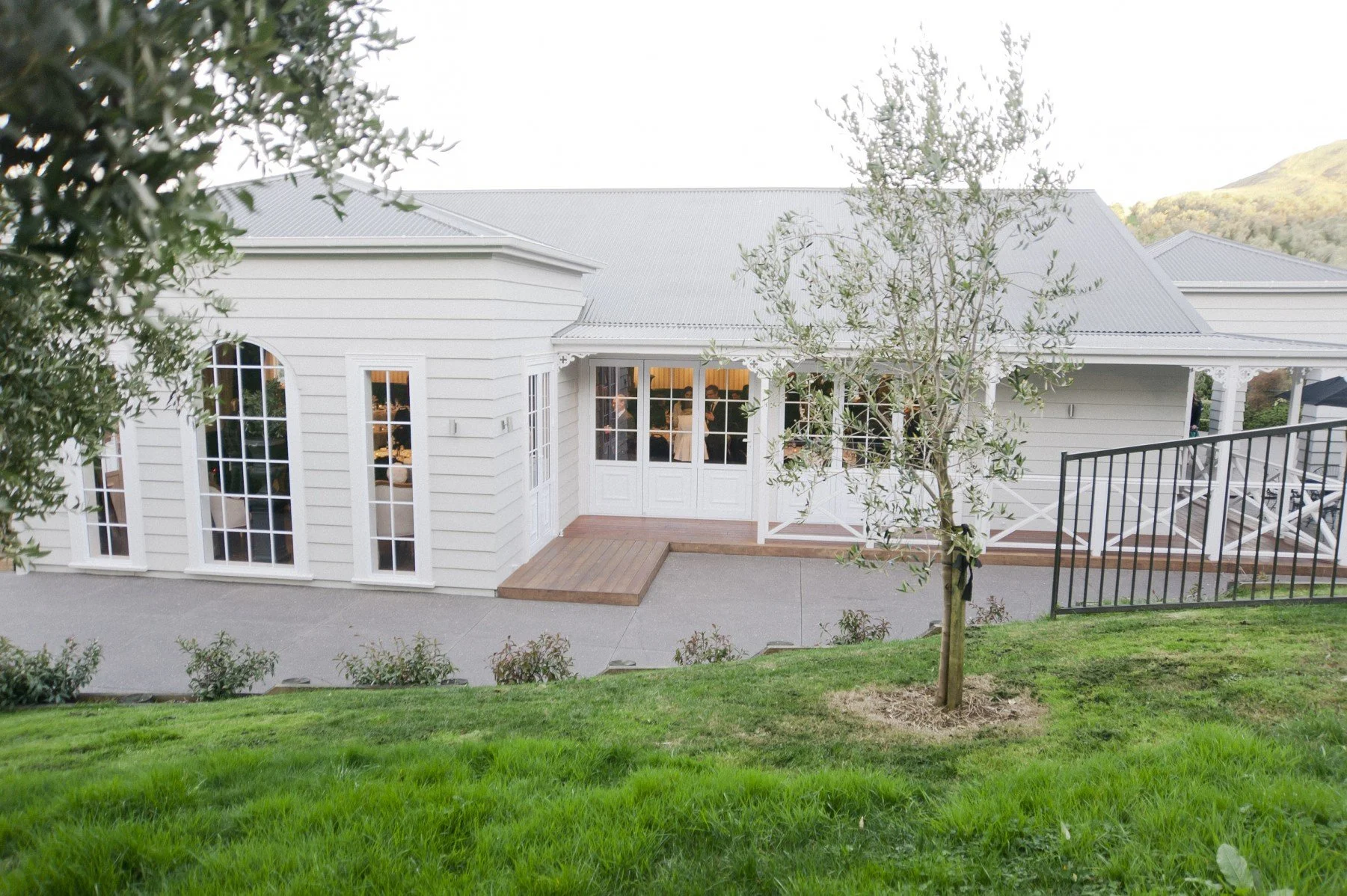 White house with large windows, a small wooden deck, and a grassy lawn with a young tree.