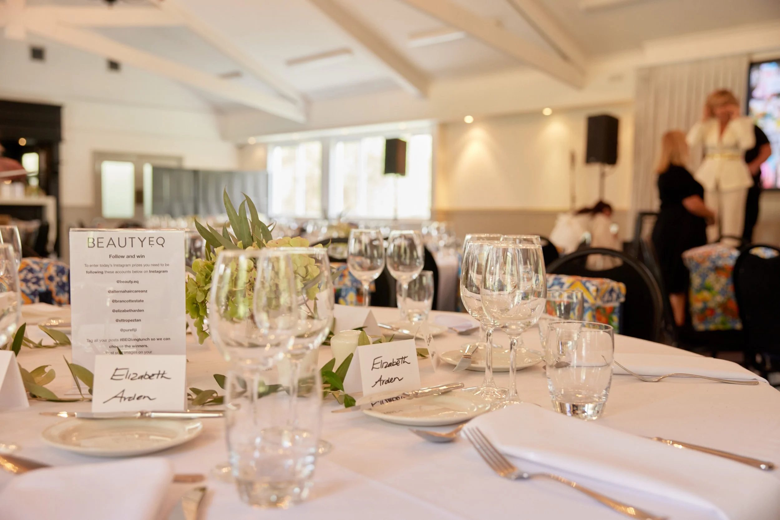 A beautifully set banquet table with glassware, plates, cutlery, and place cards, in a well-lit event space with several people and colorful chairs in the background.