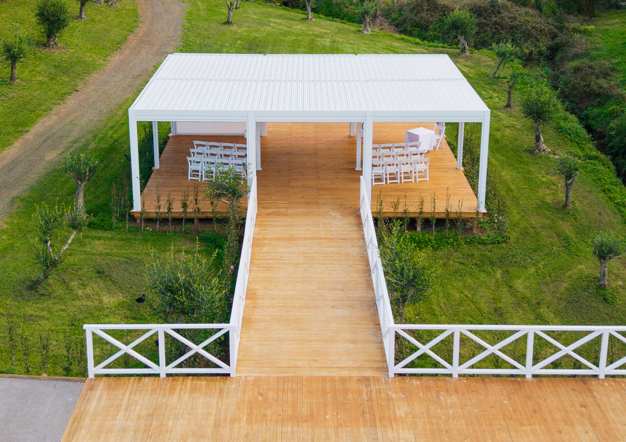 A white outdoor pavilion with a metal roof and a wooden deck, featuring white chairs and tables, surrounded by a grassy area with small trees.
