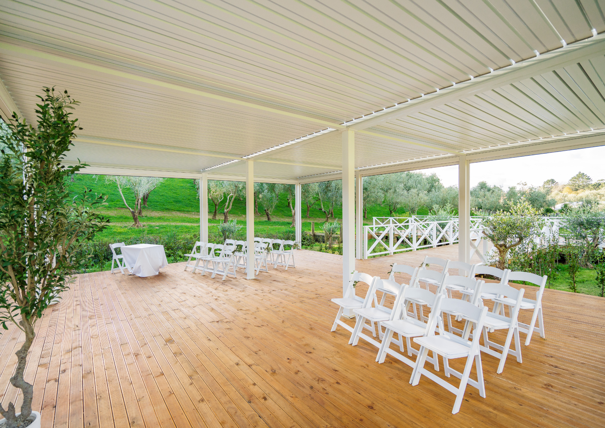 Outdoor patio with wooden floor, white folding chairs arranged in rows, a small table with a white tablecloth, surrounded by green trees and grass, with a white railing and covered ceiling.