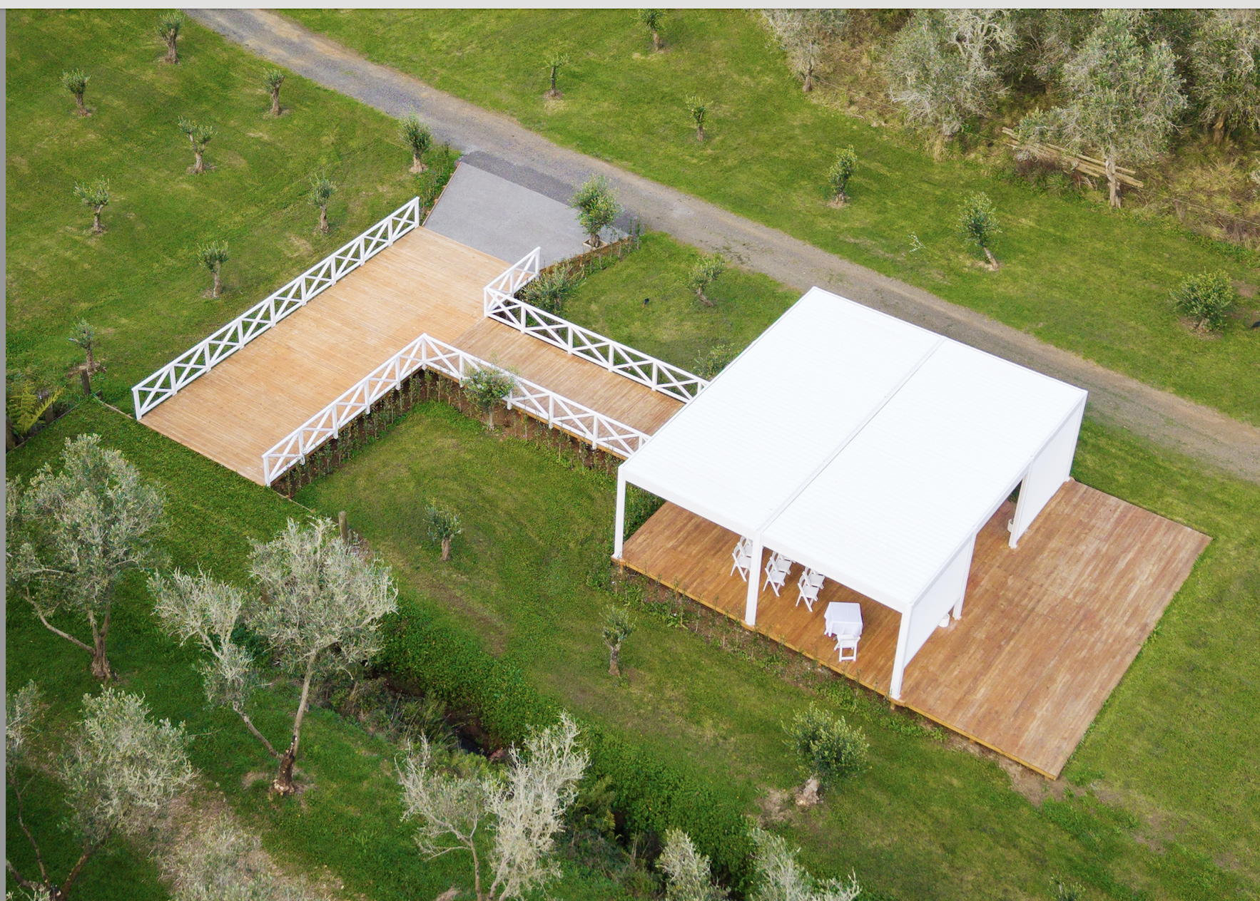 Aerial view of a backyard with a wooden deck attached to a white building, surrounded by green grass, trees, and a gravel pathway.