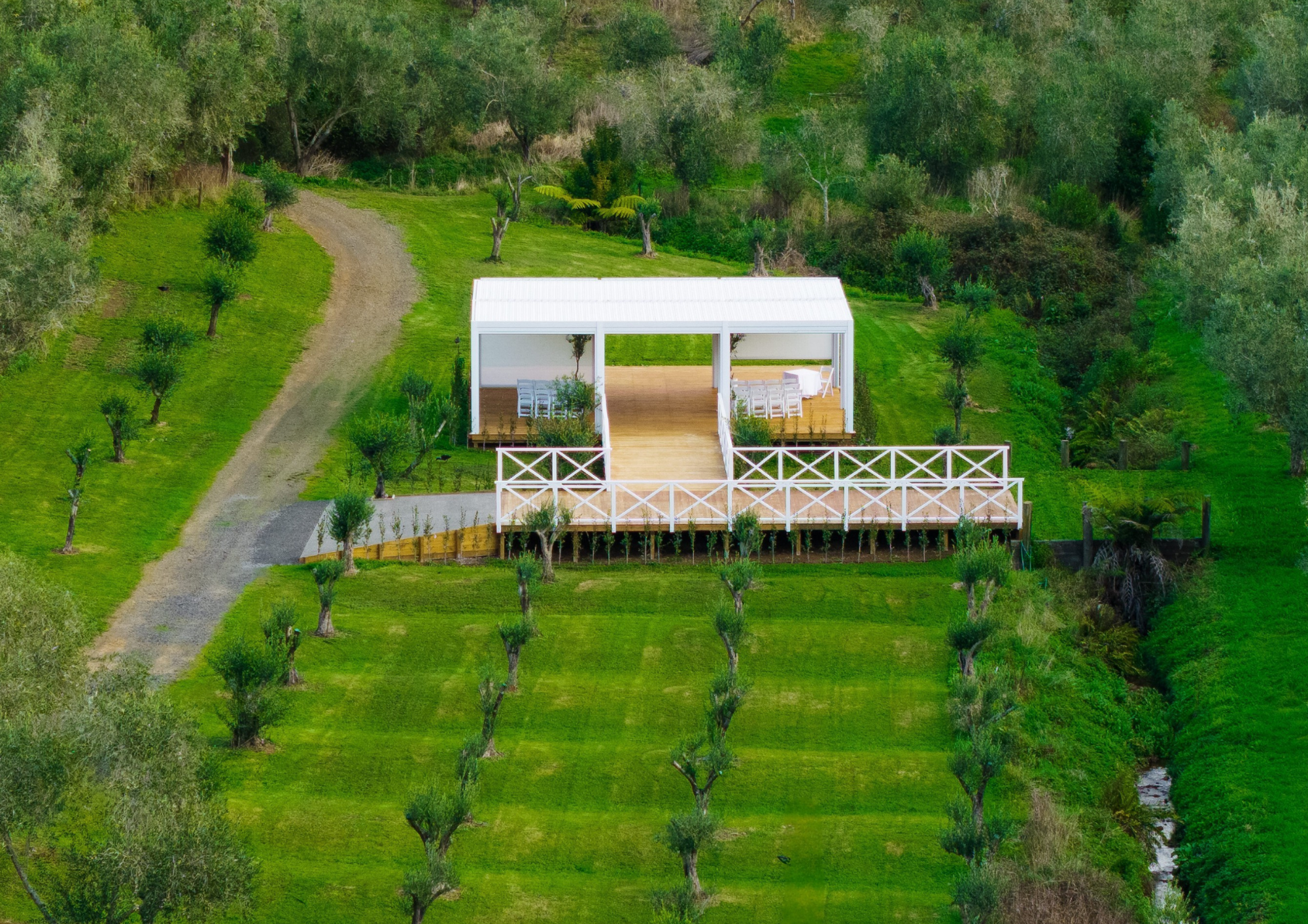 A white outdoor pavilion with a wooden floor, surrounded by trees and grass, with a dirt path leading up to it.