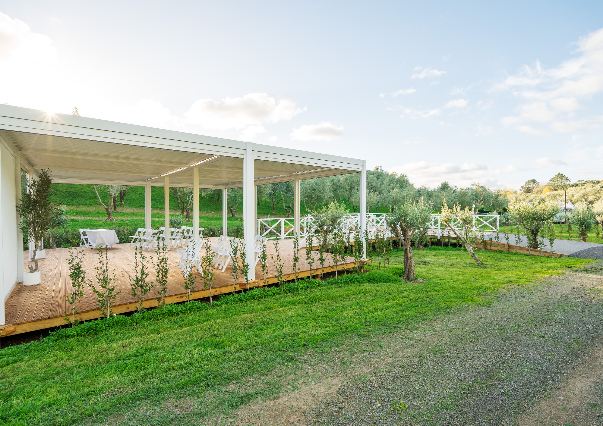 Outdoor event space with a wooden deck, white chairs, small round tables with white tablecloths, potted trees, and greenery, set against a lush green landscape with a partly cloudy sky.