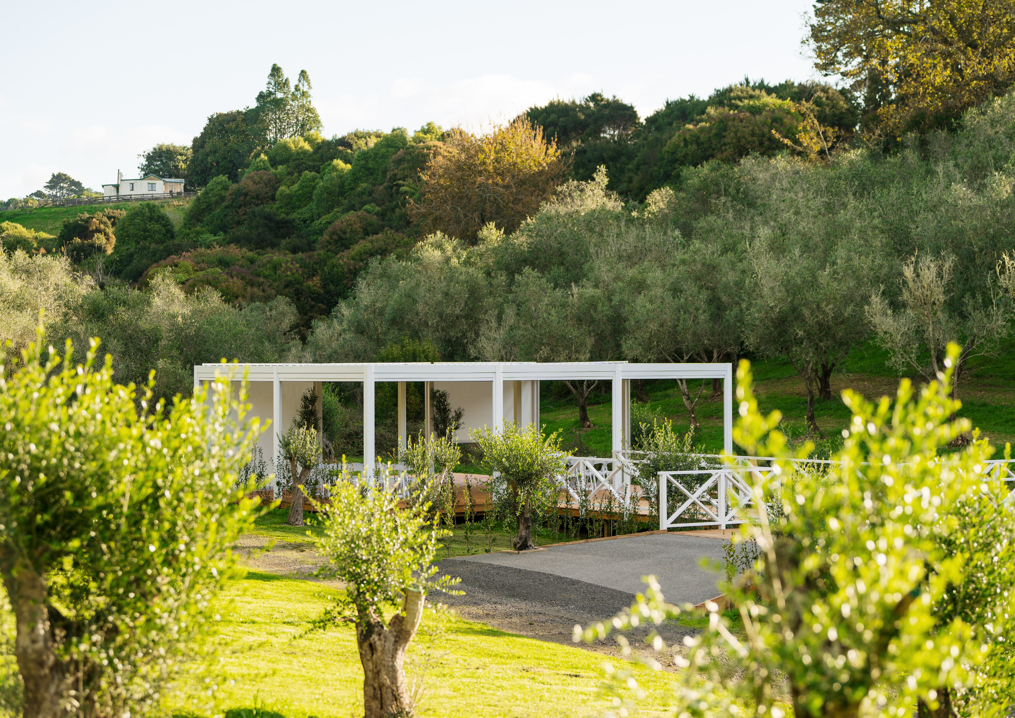 A modern white structure with a deck, surrounded by trees and green hills, under a partly cloudy sky.