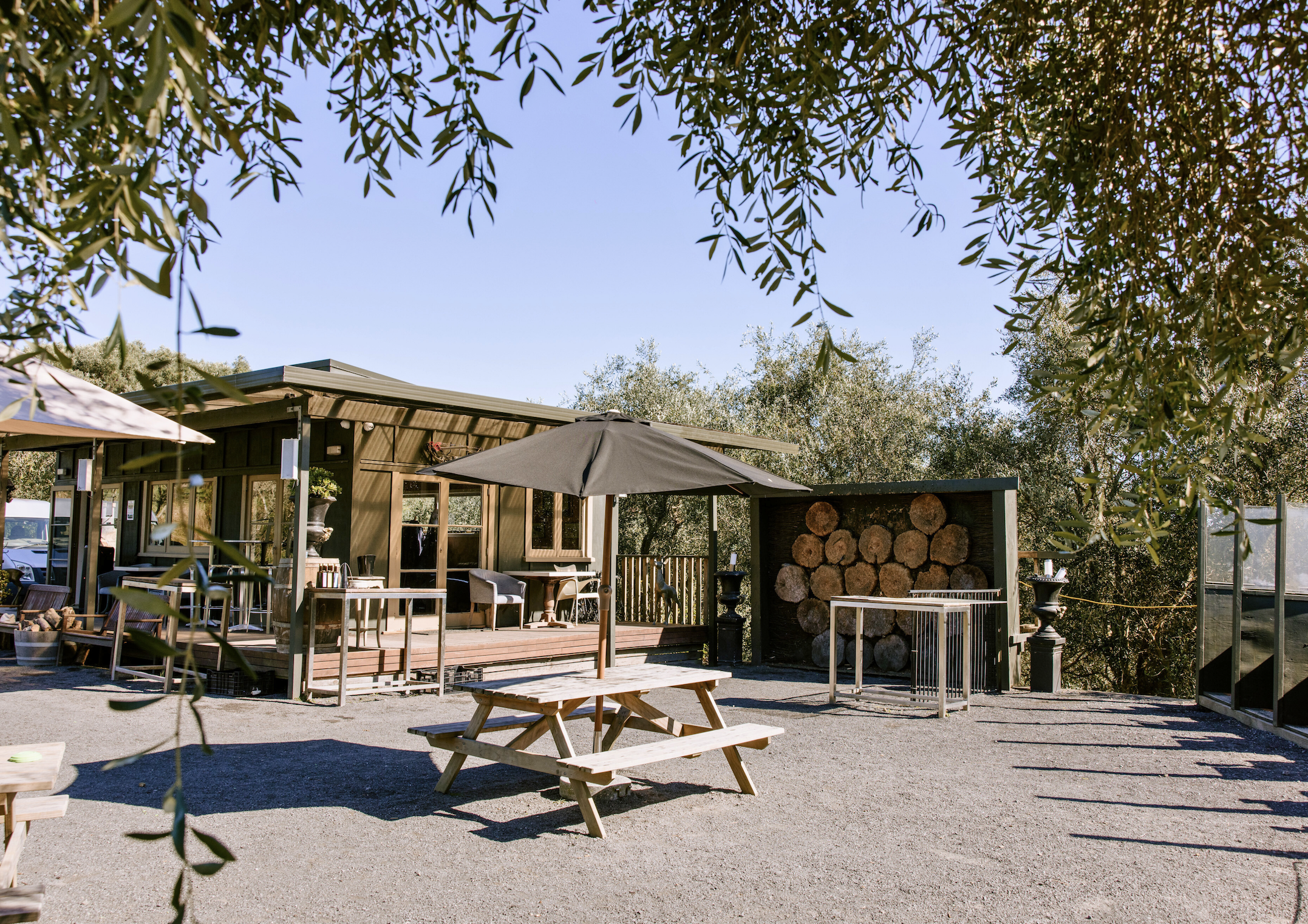 Outdoor patio with wooden picnic table, patio umbrella, wooden logs stacked against a wall, and a small building with a porch, greenery, and bright blue sky.