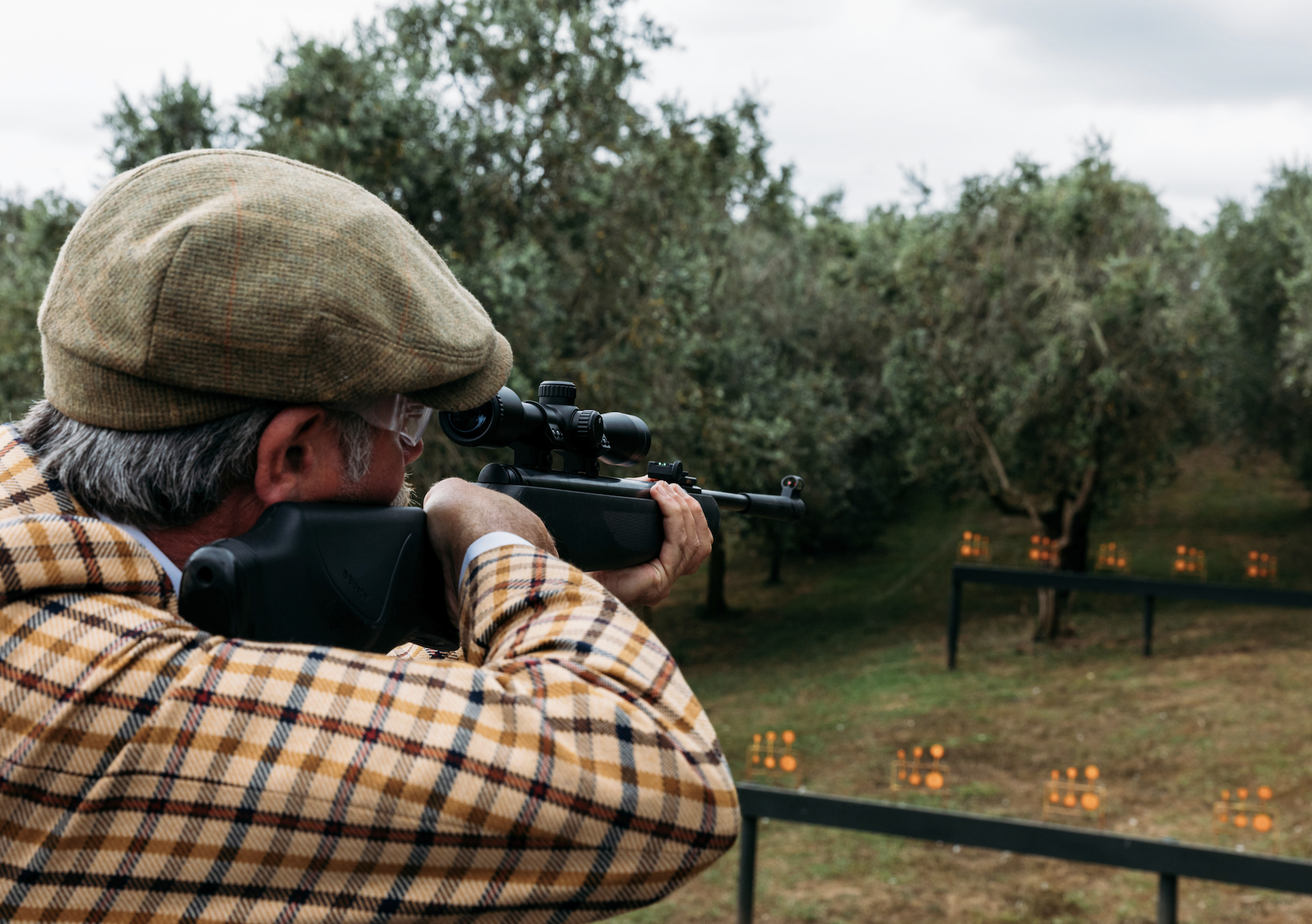 Man aiming with a rifle at outdoor shooting range with targets and trees in the background.