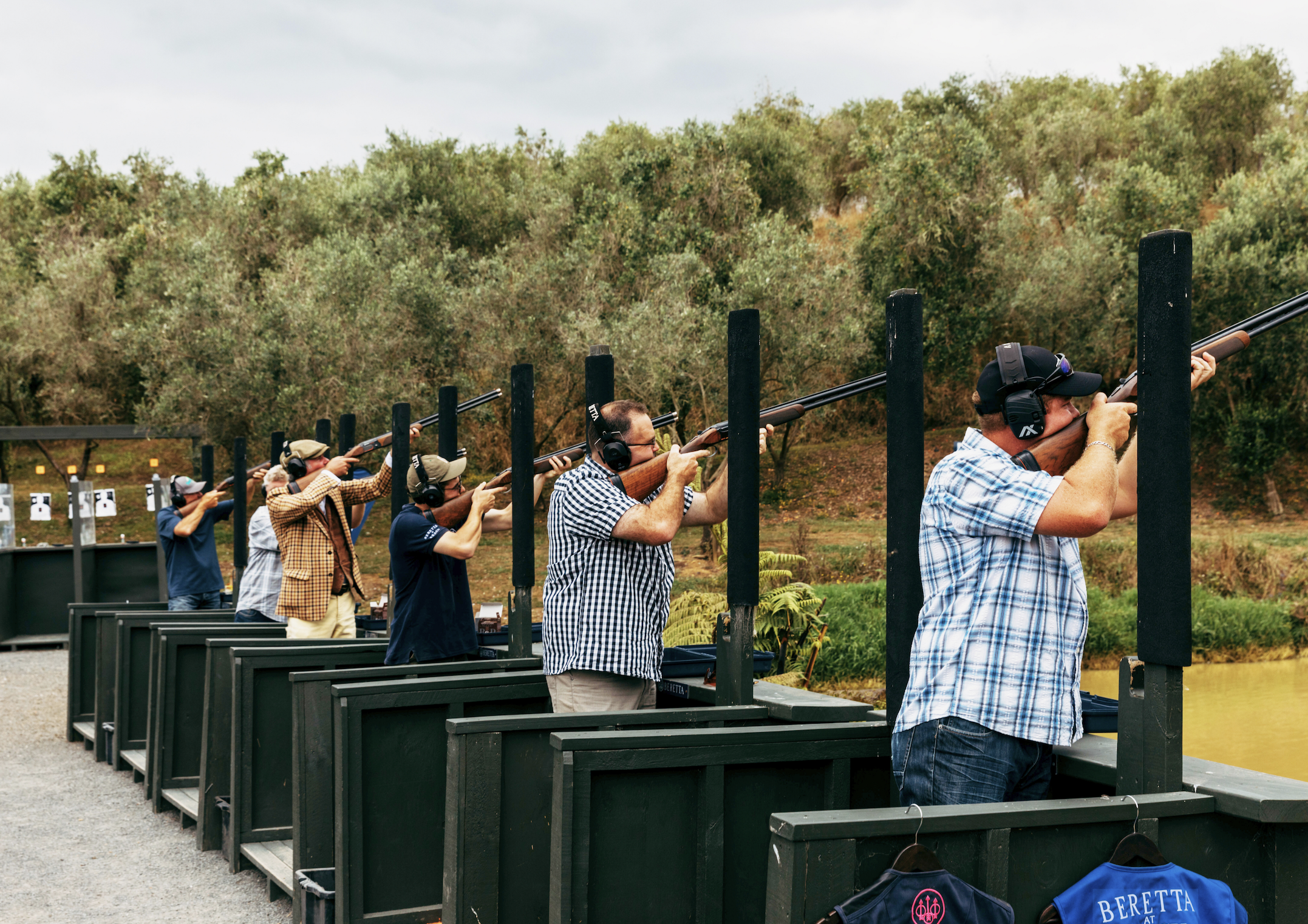Group of people at outdoor shooting range aiming rifles at targets, wearing ear protection and casual clothing.