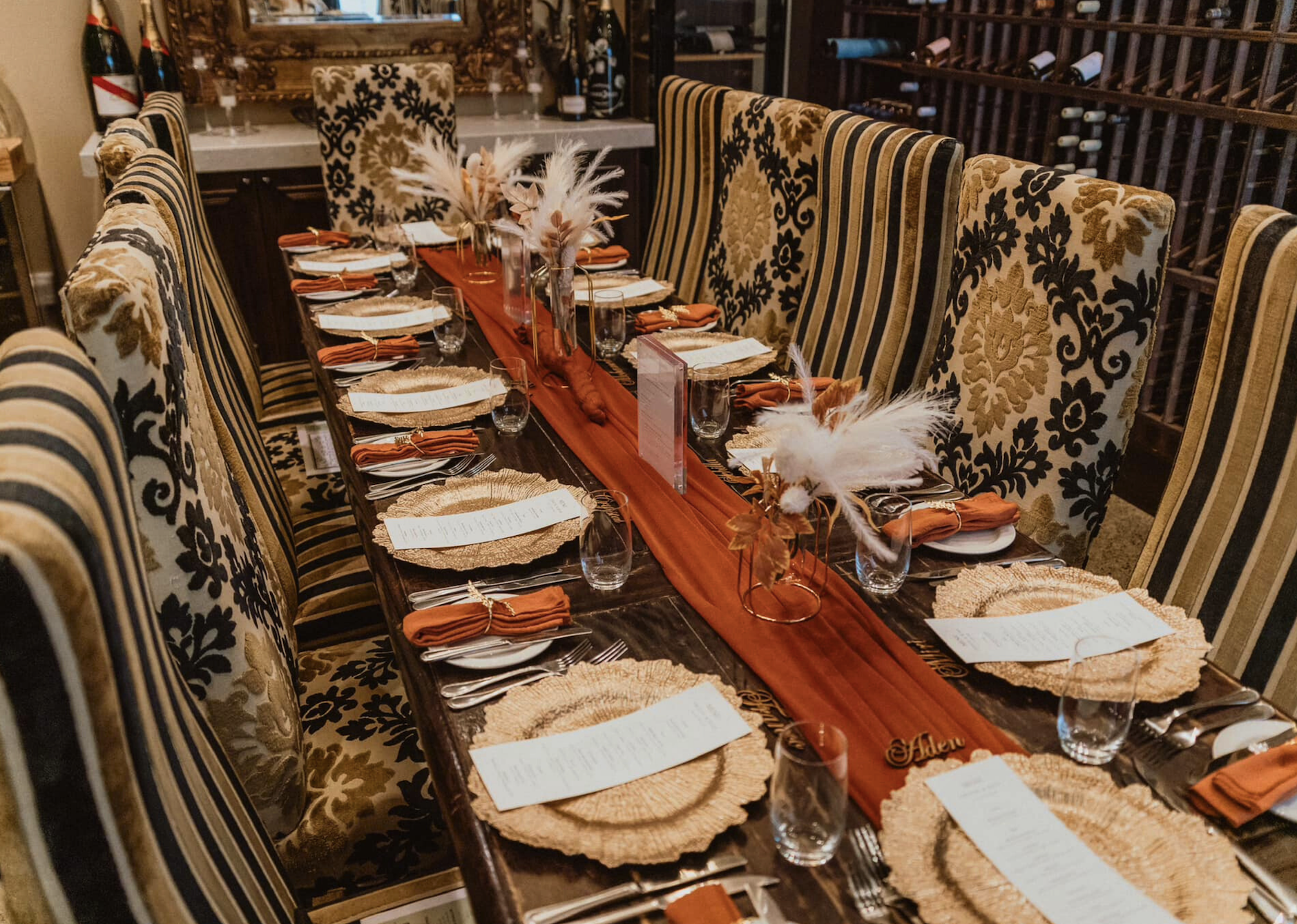 A long wooden dining table set for a formal meal with orange napkins, woven placemats, and clear glassware. The table has tall chairs with black and gold floral patterns and is decorated with a burnt orange table runner, feathered centerpieces, and name tags.