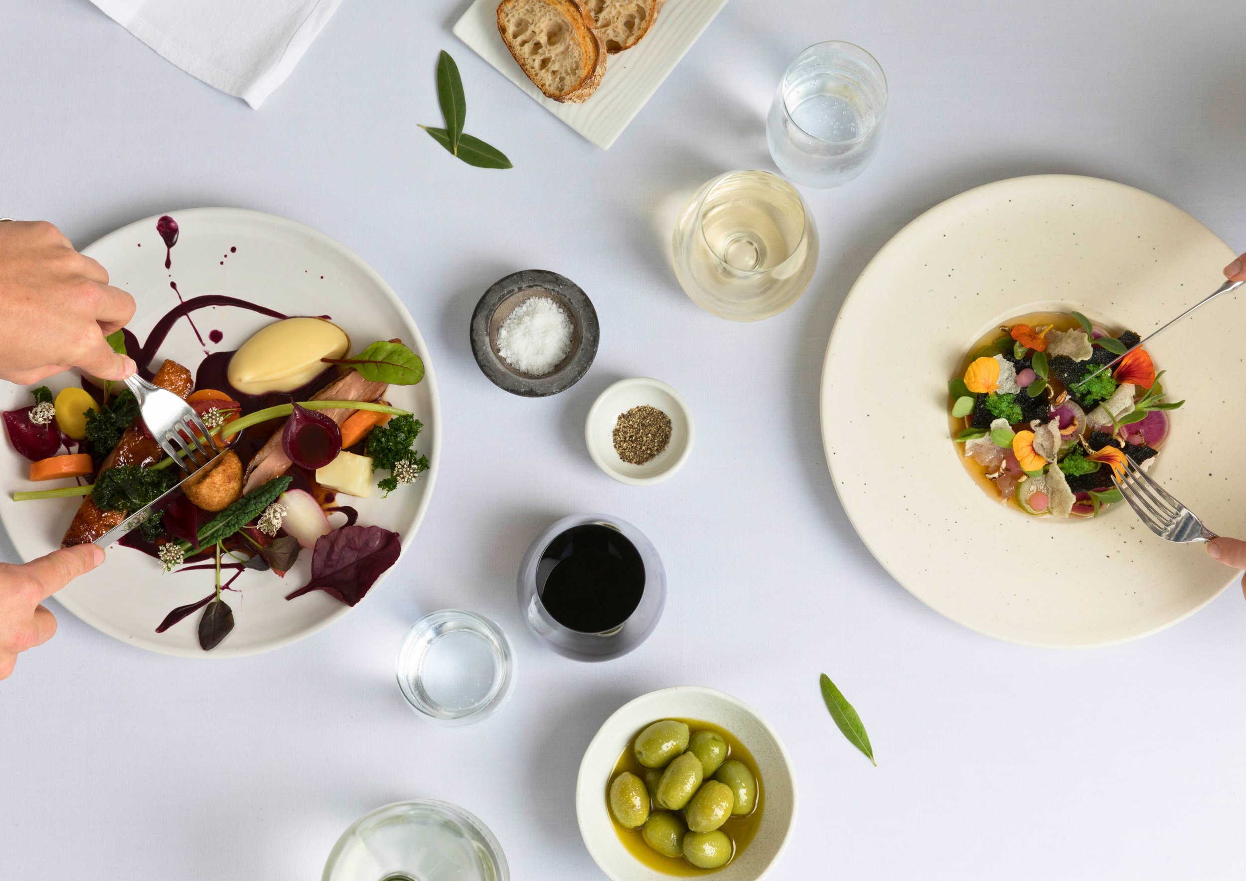 Overhead photo of a fine dining table with two plates of colorful salads, various glasses of beverages, small bowls of olives, coarse salt, and spices, with a plate of bread slices at the top.