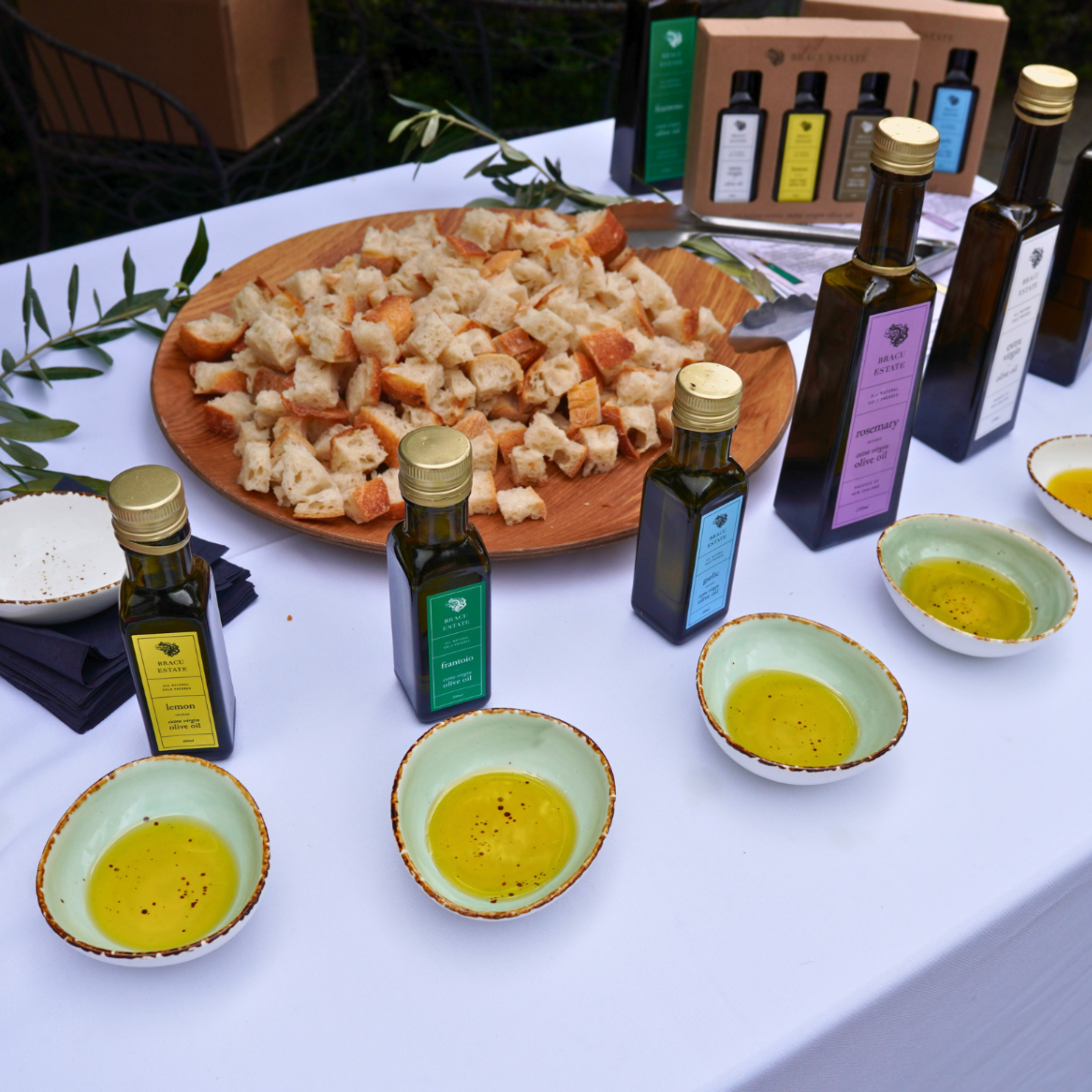 Table with bowls of olive oil, bottles of flavored olive oils, a plate of bread cubes, and olive leaves, likely for a tasting event.