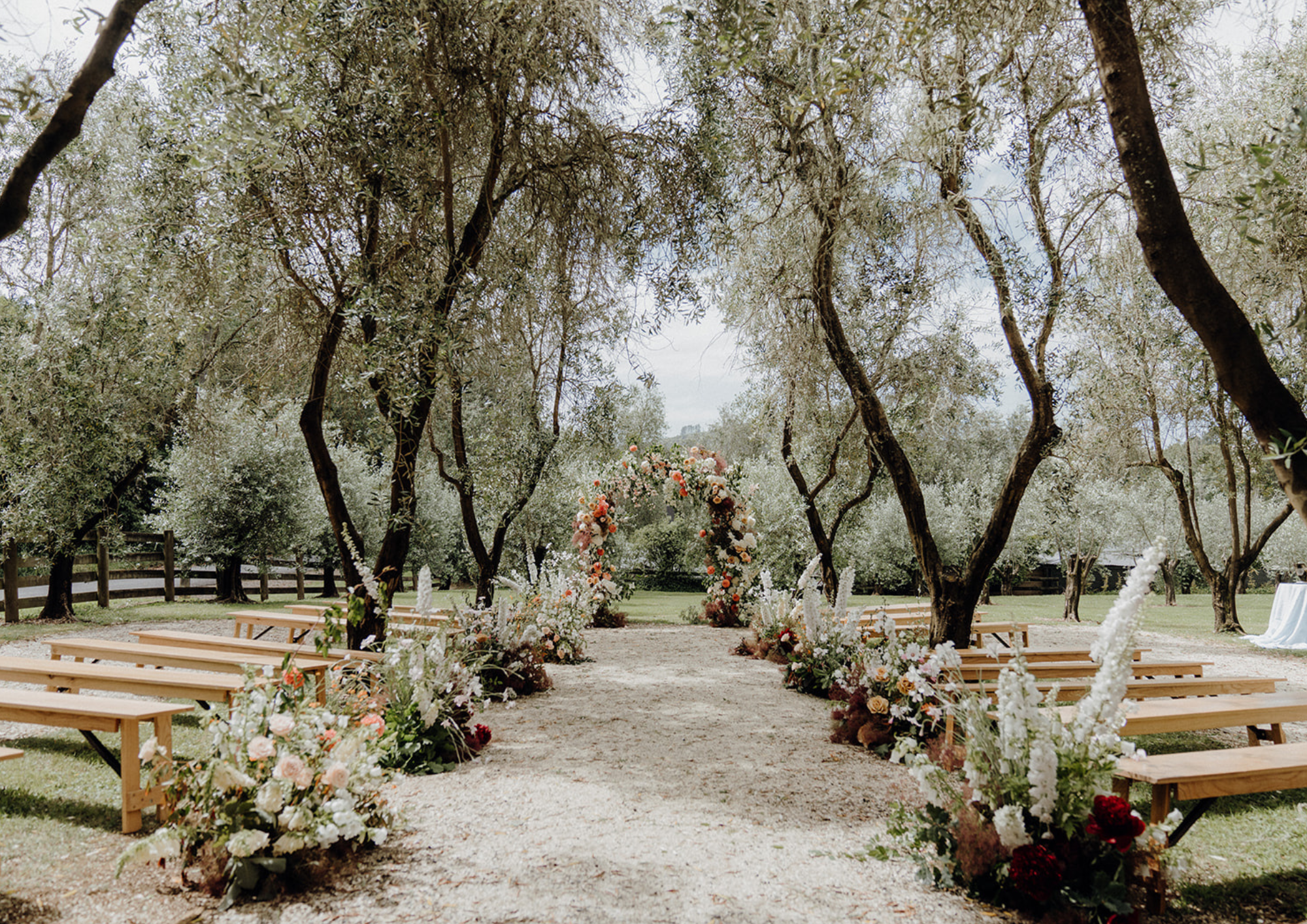 Outdoor wedding ceremony setup with wooden benches, floral archway, and decorative flower arrangements along the pathway, under a canopy of trees in a natural setting.