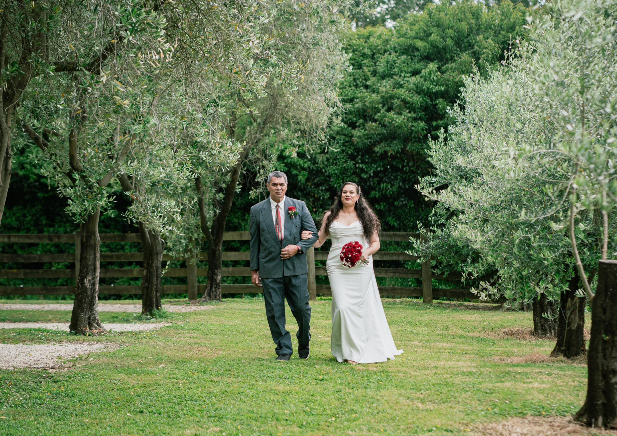 A bride in a white wedding dress walking down a grassy pathway with an older man in a suit, holding her arm, surrounded by green trees and a wooden fence.