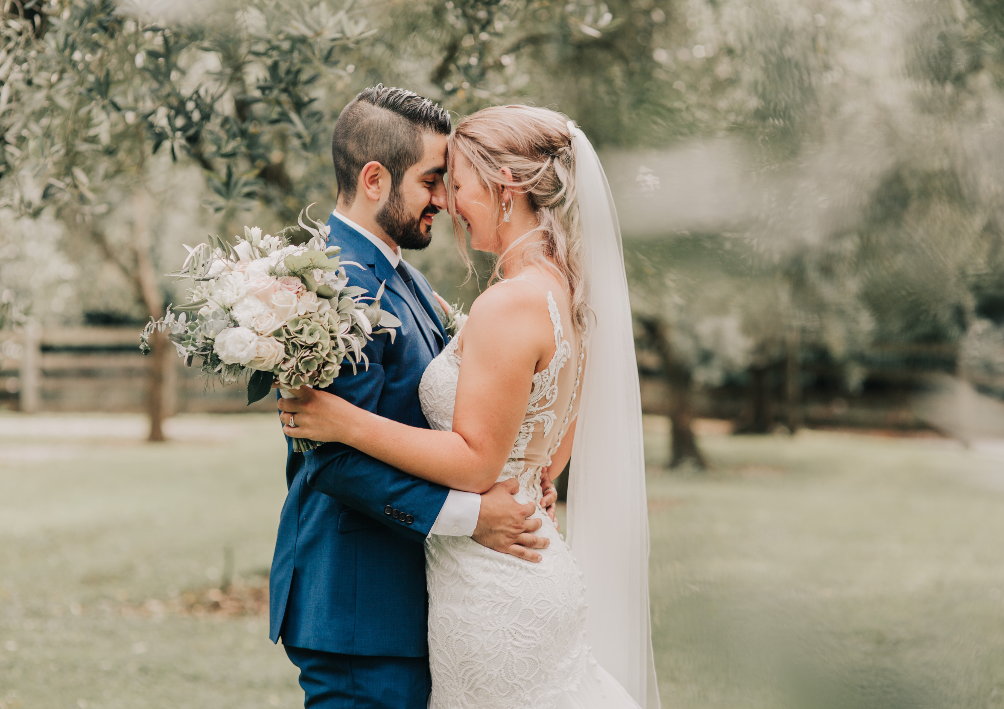 A bride and groom embrace closely outdoors, foreheads touching, with eyes closed, smiling softly. The bride holds a large bouquet of white and pink flowers with greenery, and wears a lace wedding dress with a veil. The groom wears a blue suit with a white shirt and tie. The background features blurred trees and grass, creating a romantic, natural setting.