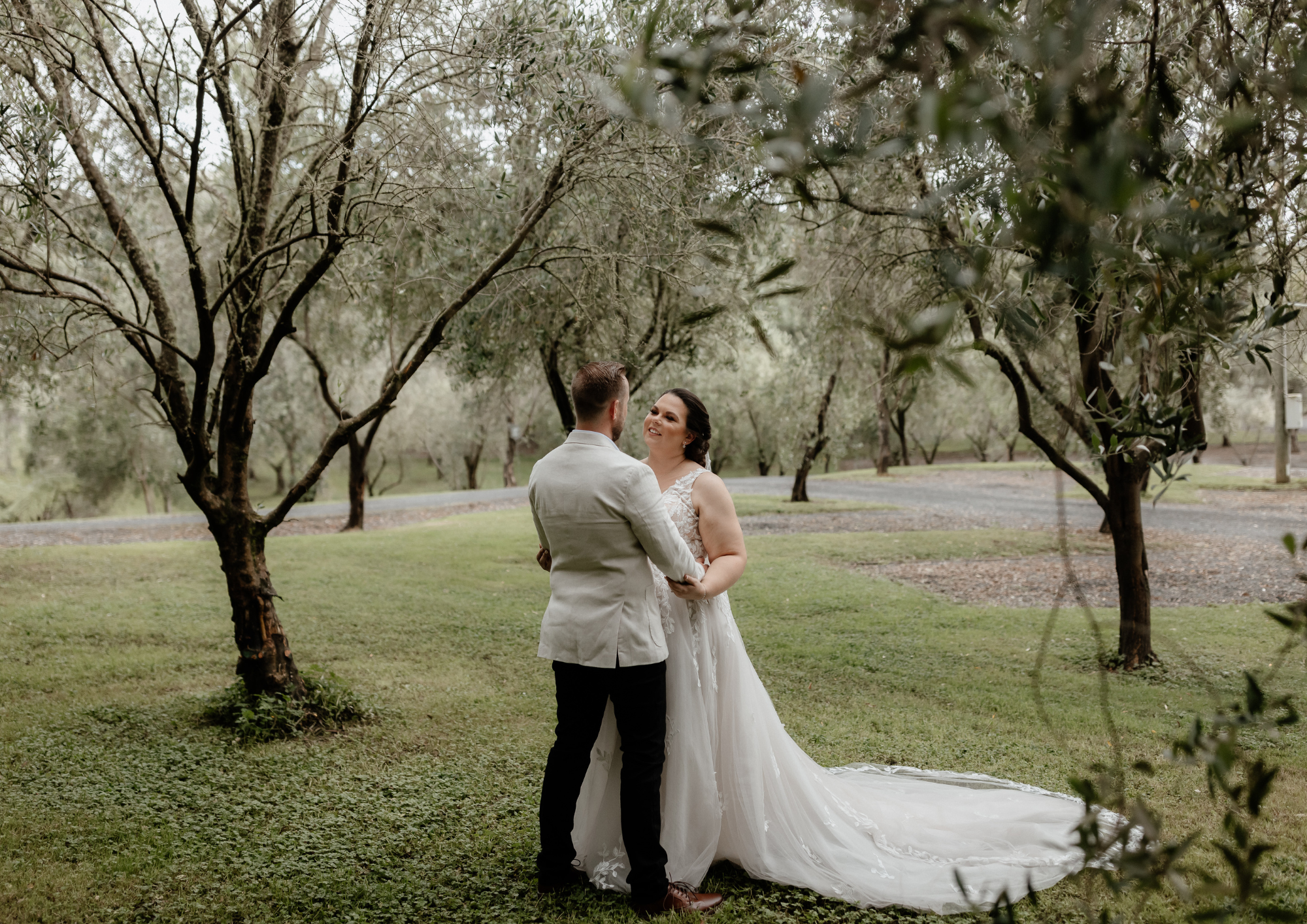 A bride and groom standing on grass beneath trees, facing each other and smiling, in an outdoor wedding setting.