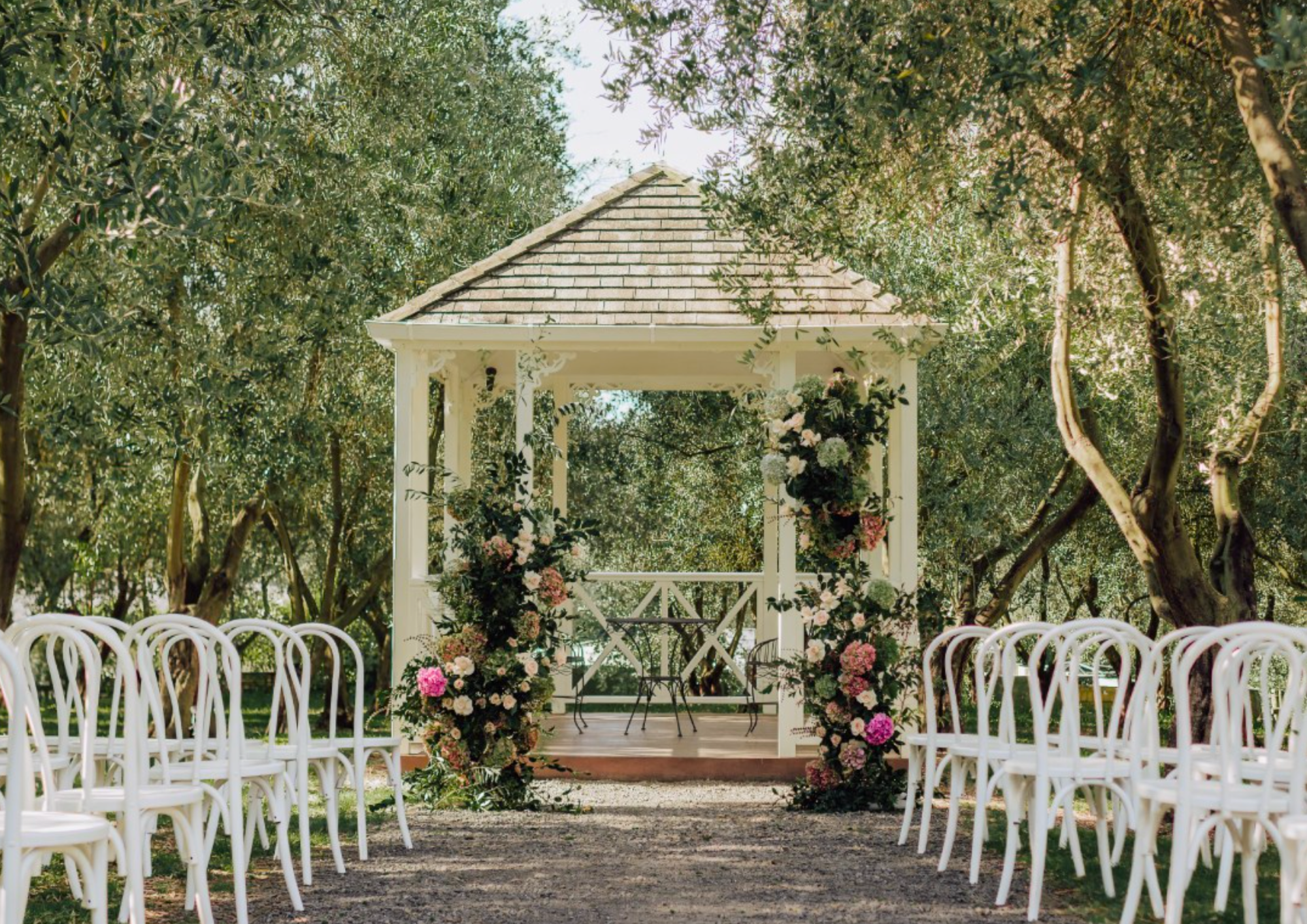 Outdoor wedding ceremony setup with white chairs facing a small white pavilion decorated with pink and white flowers, surrounded by trees.