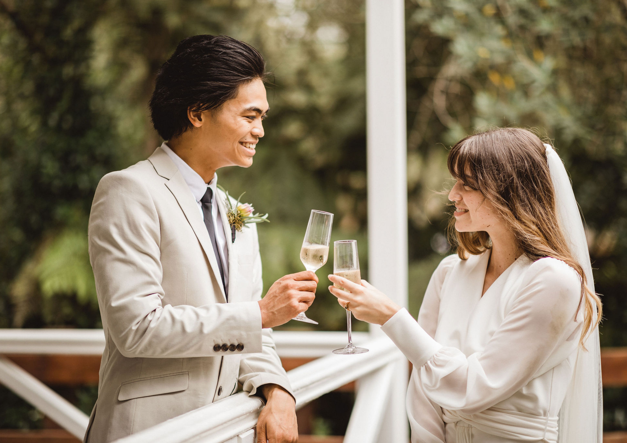 A man and a woman in formal wedding attire sharing a toast with champagne on a balcony with lush greenery in the background.