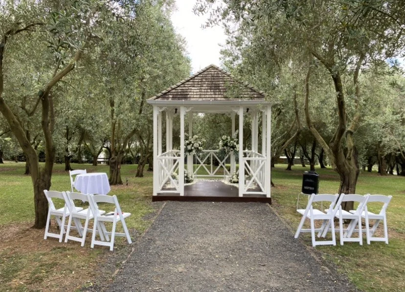 Small white gazebo decorated with flowers, set for an outdoor wedding ceremony in a wooded park with white folding chairs arranged on either side of an aisle.