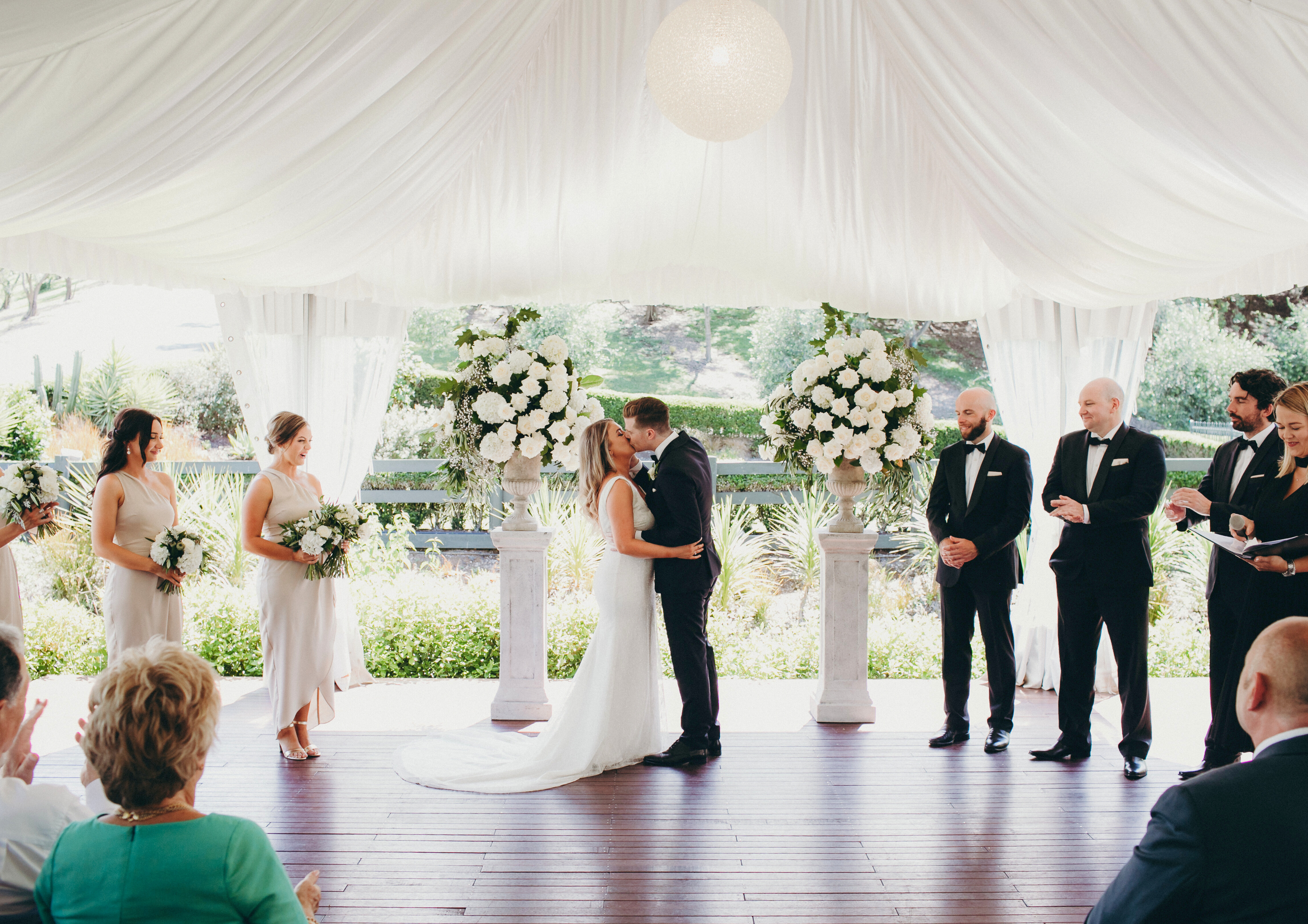 A wedding ceremony with a bride and groom kissing under a white tent. Bridesmaids and groomsmen stand on either side, with floral decorations and guests watching.