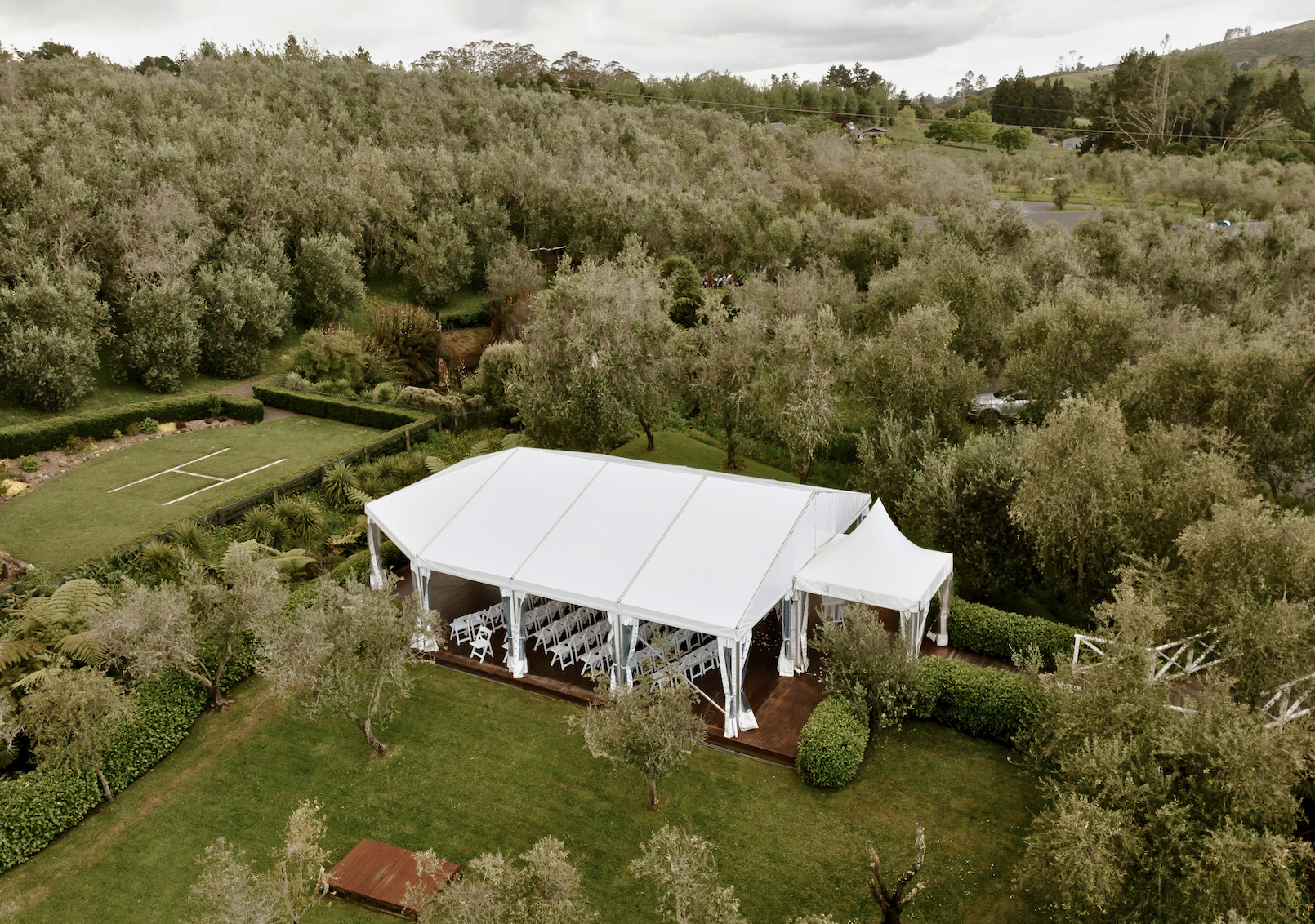 Large white event tent set up on a grassy area surrounded by trees and shrubs in a lush outdoor setting.