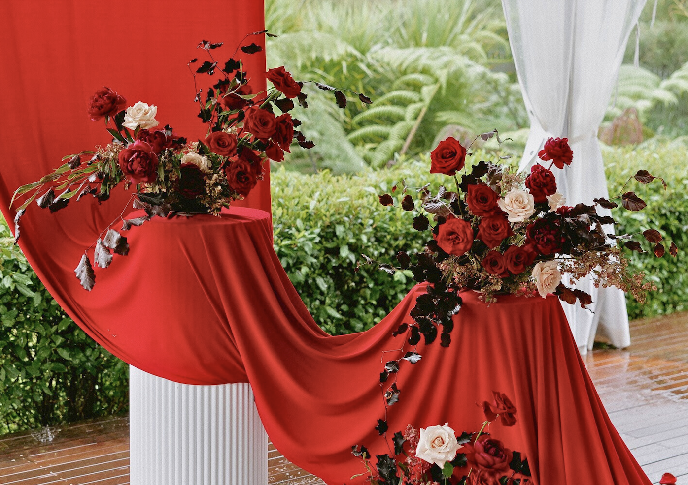 Two floral arrangements with red and cream roses and dark foliage placed on red fabric-draped tables with a white column, outside on a wooden deck with greenery and hills in the background.