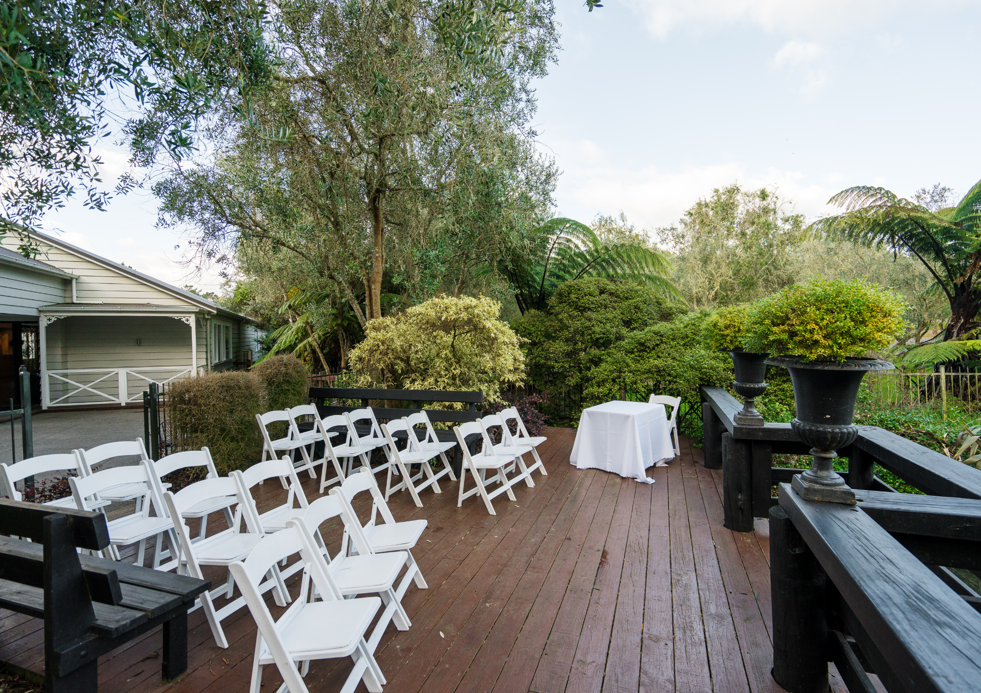 Outdoor wedding ceremony setup on a wooden deck with white folding chairs arranged in rows and a small table with a white tablecloth, surrounded by lush green trees and plants, under a partly cloudy sky.