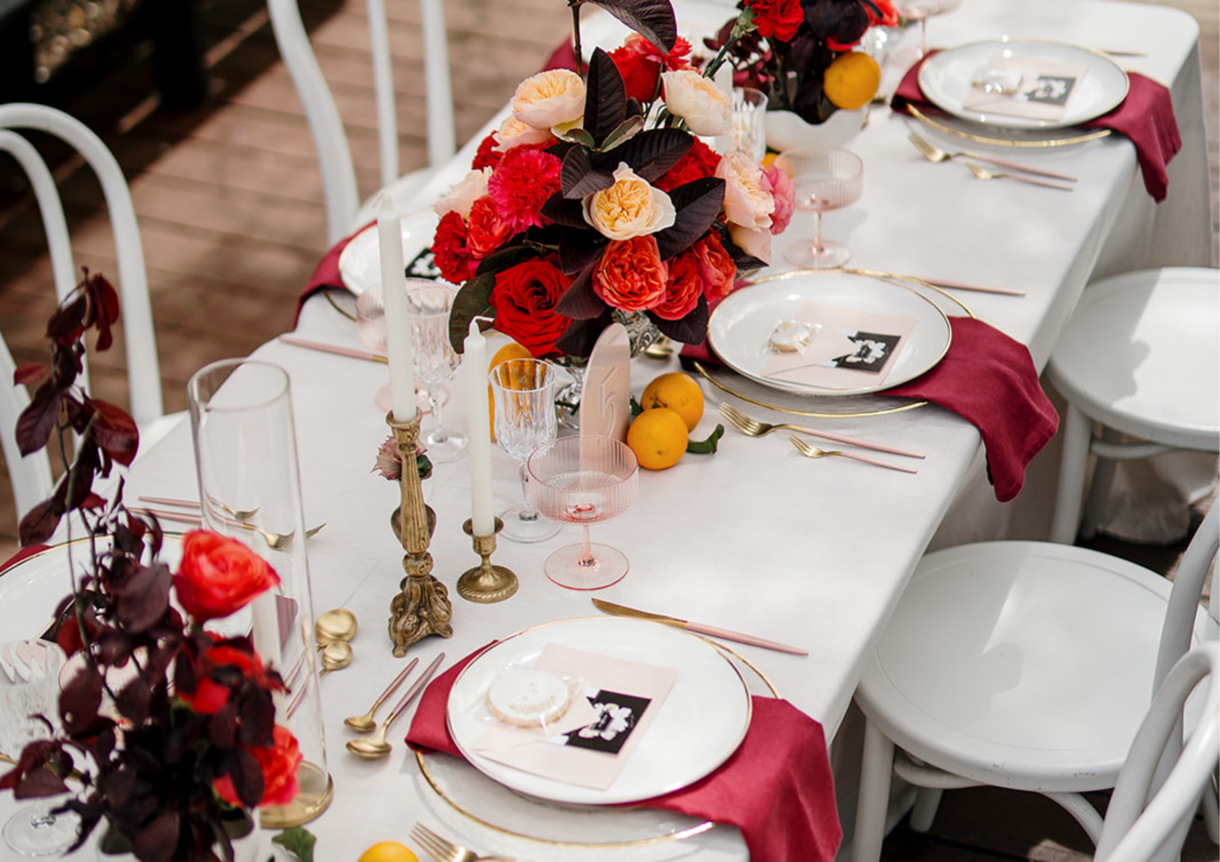 A decorated dining table with a white tablecloth, a large floral centerpiece with red, pink, peach, and dark purple flowers, gold and pink glassware, gold cutlery, cream-colored plates with black accents, burgundy napkins, tall white candles in gold holders, and small yellow decorative fruits.