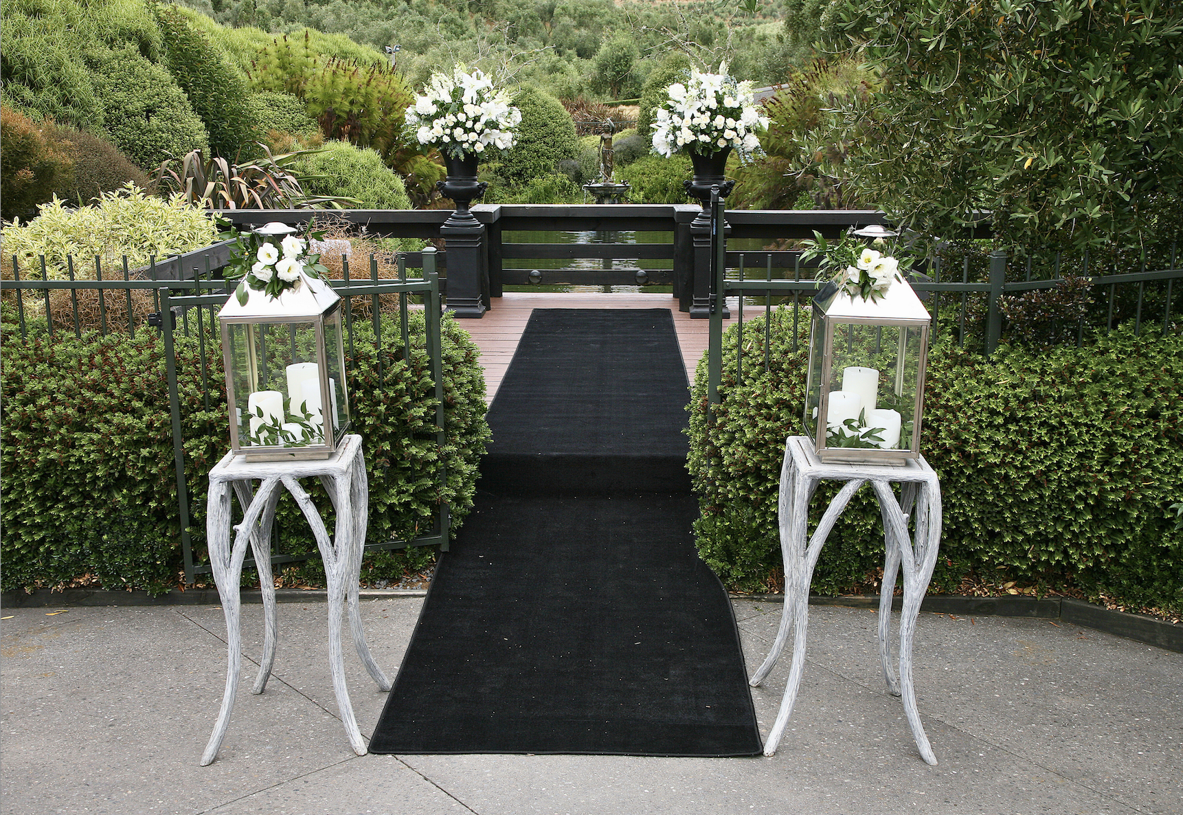 Decorative pathway with lanterns and white flowers leading to a small bridge over a pond, surrounded by lush green bushes and trees.