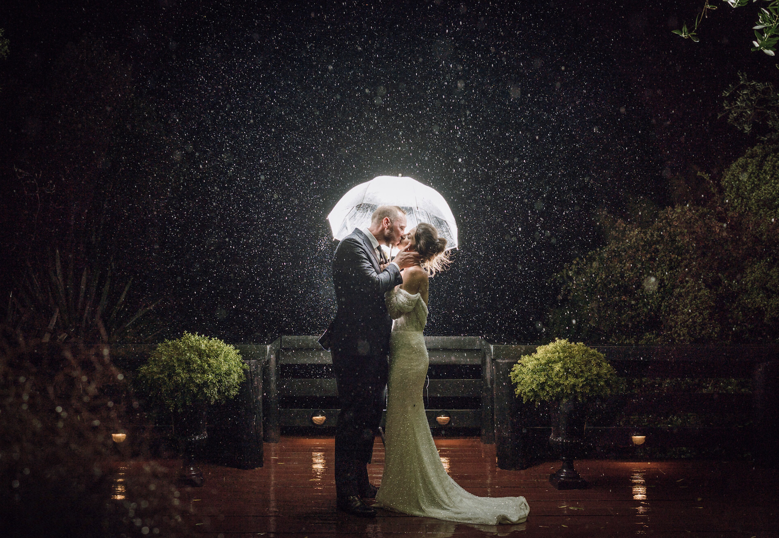 A bride and groom sharing a kiss under an umbrella on a rainy night, illuminated from behind, with raindrops falling around them and potted bushes on a wooden deck.