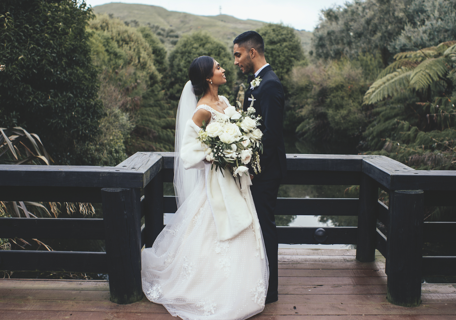A bride and groom standing close on a wooden bridge with a lush green landscape and river in the background, celebrating their wedding.