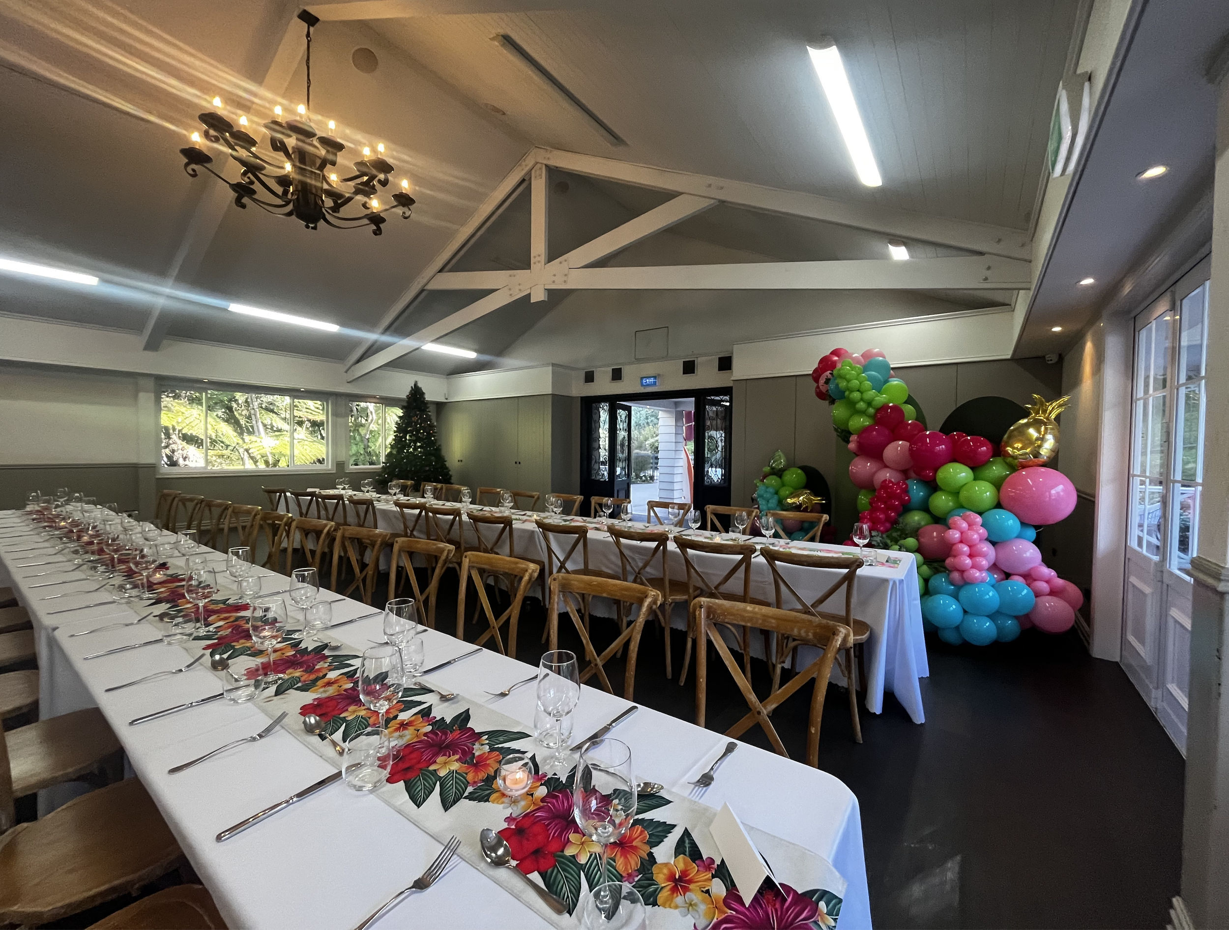 Banquet hall decorated for a celebration with long tables, floral table runners, and colorful balloon arrangements including a gold pineapple balloon. There is a Christmas tree in the corner and the room has large windows and a chandelier.