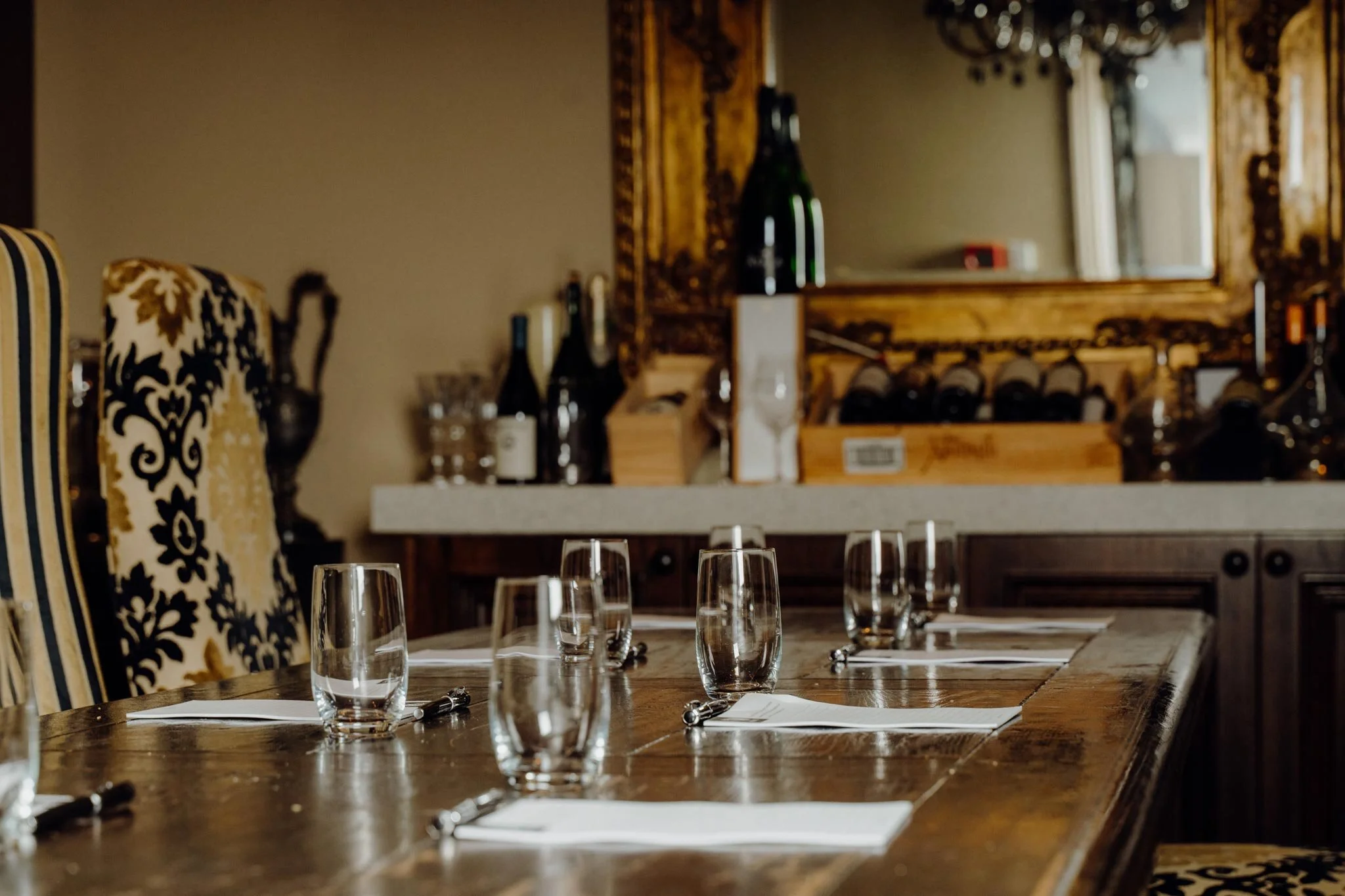 A dining table set with water glasses, silverware, and napkins in a restaurant with ornate chairs, bottles, and a large mirror in the background.