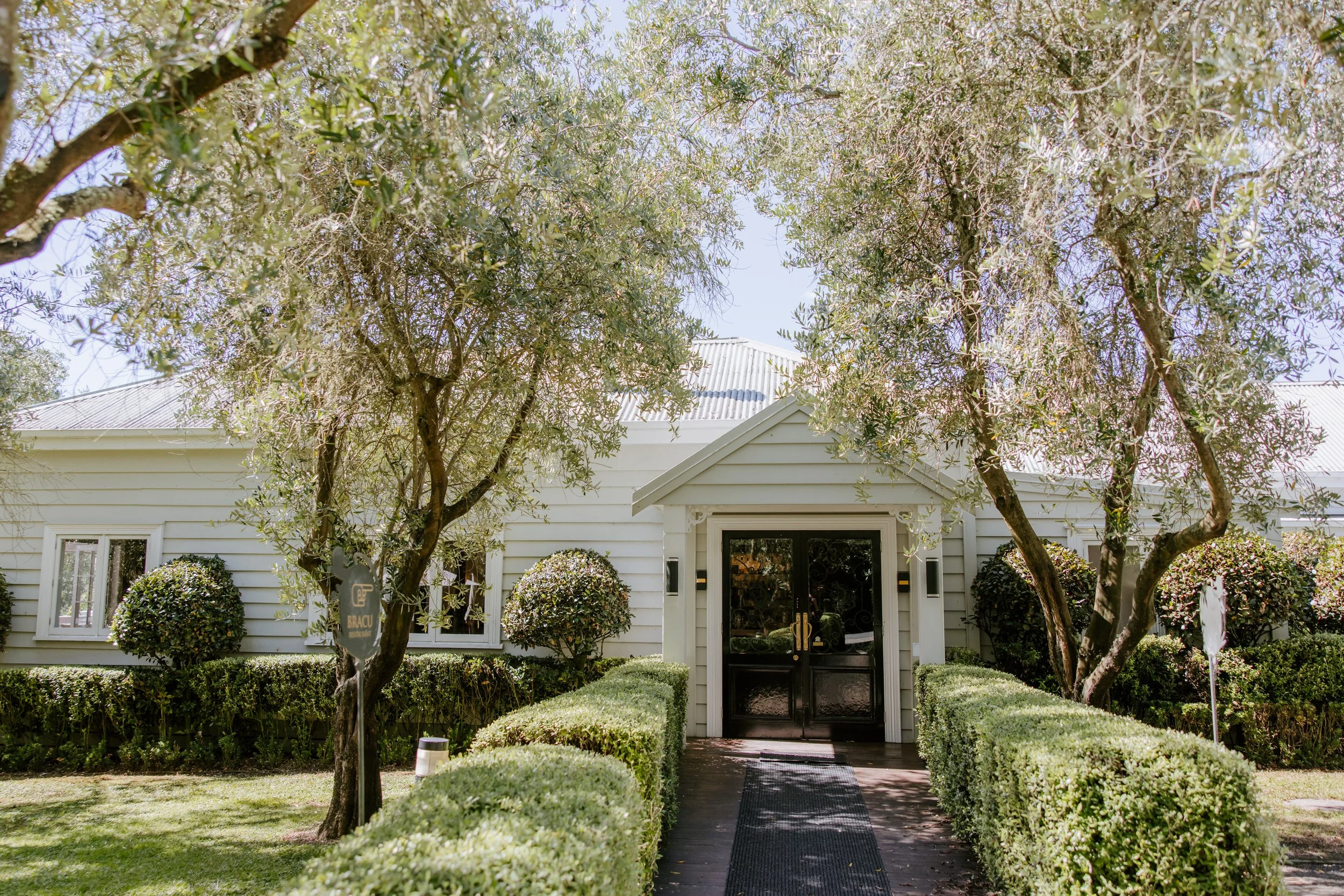 White house with black doors, surrounded by neatly trimmed bushes and trees, on a sunny day.