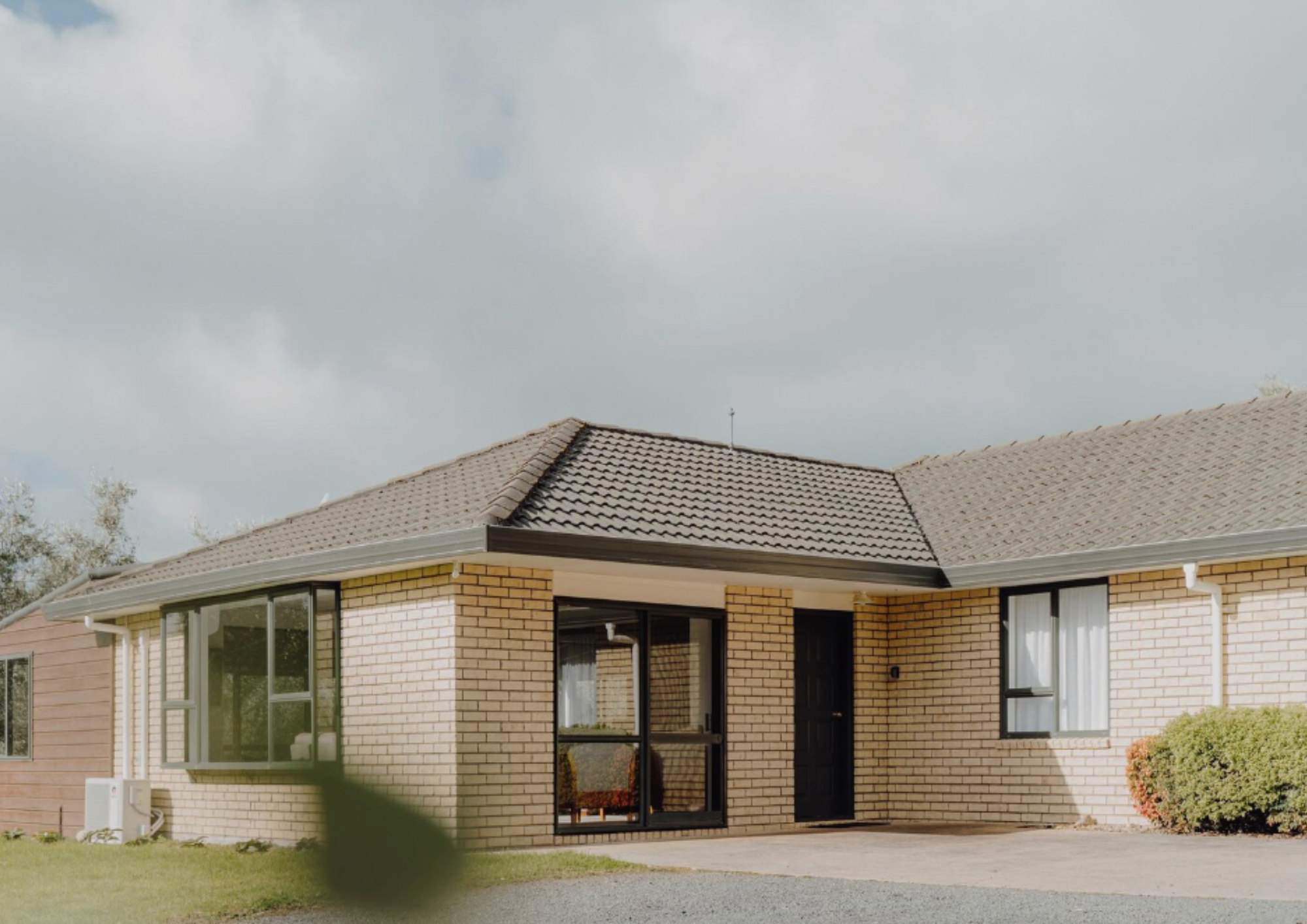 A single-story brick house with a dark front door, large windows, and a sloped tiled roof, surrounded by a small lawn and bushes.