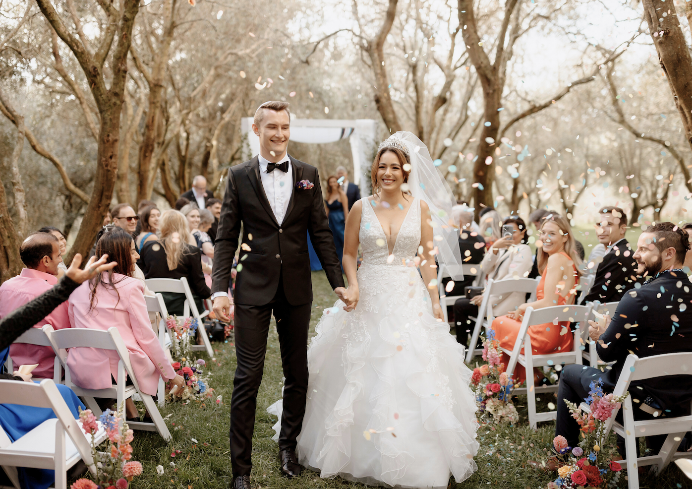 A newlywed couple walking hand-in-hand down the aisle at an outdoor wedding ceremony, surrounded by friends and family, with confetti falling around them in a wooded setting.