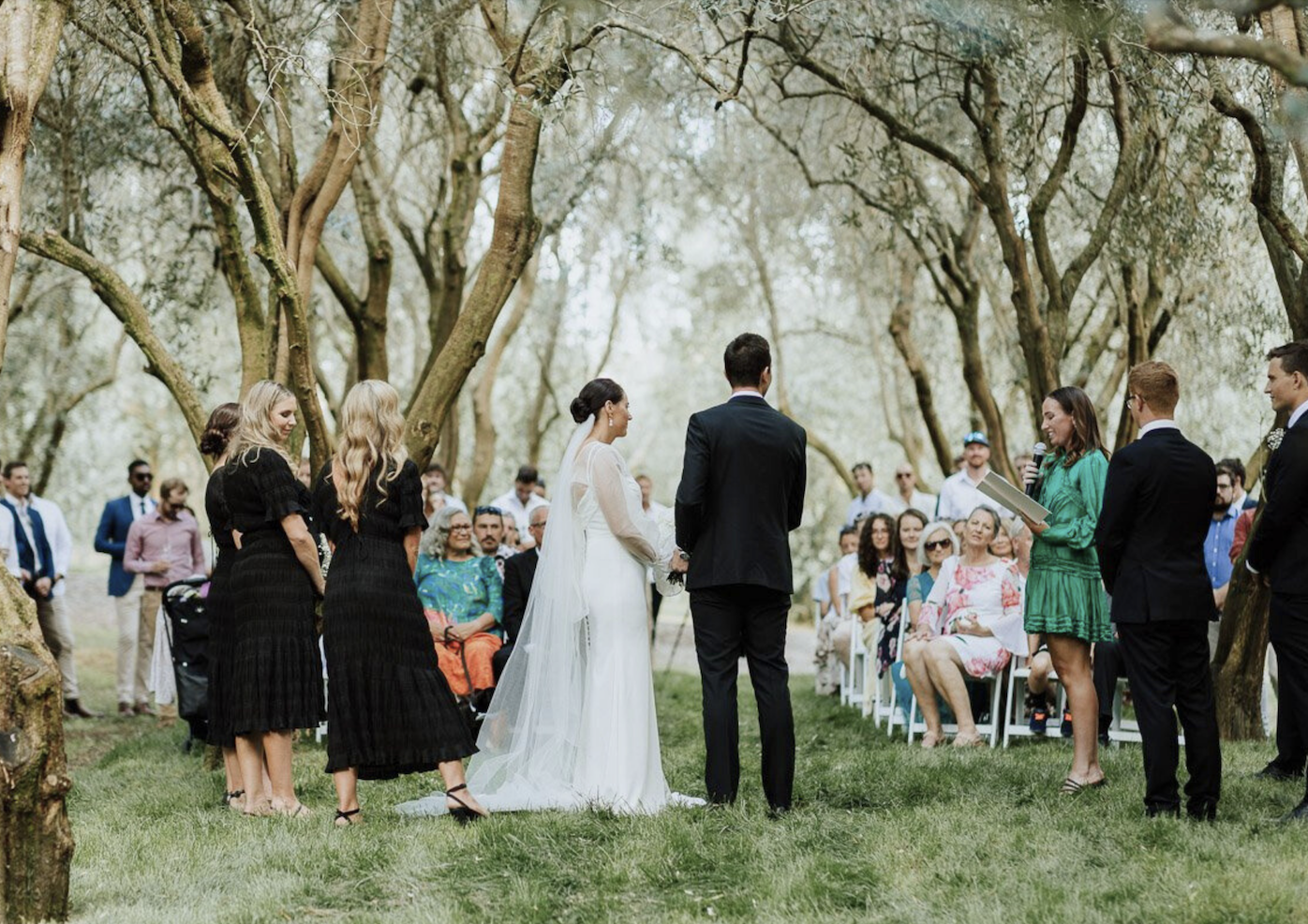 A wedding ceremony outdoors under trees with the bride and groom standing before an officiant and guests seated in rows.