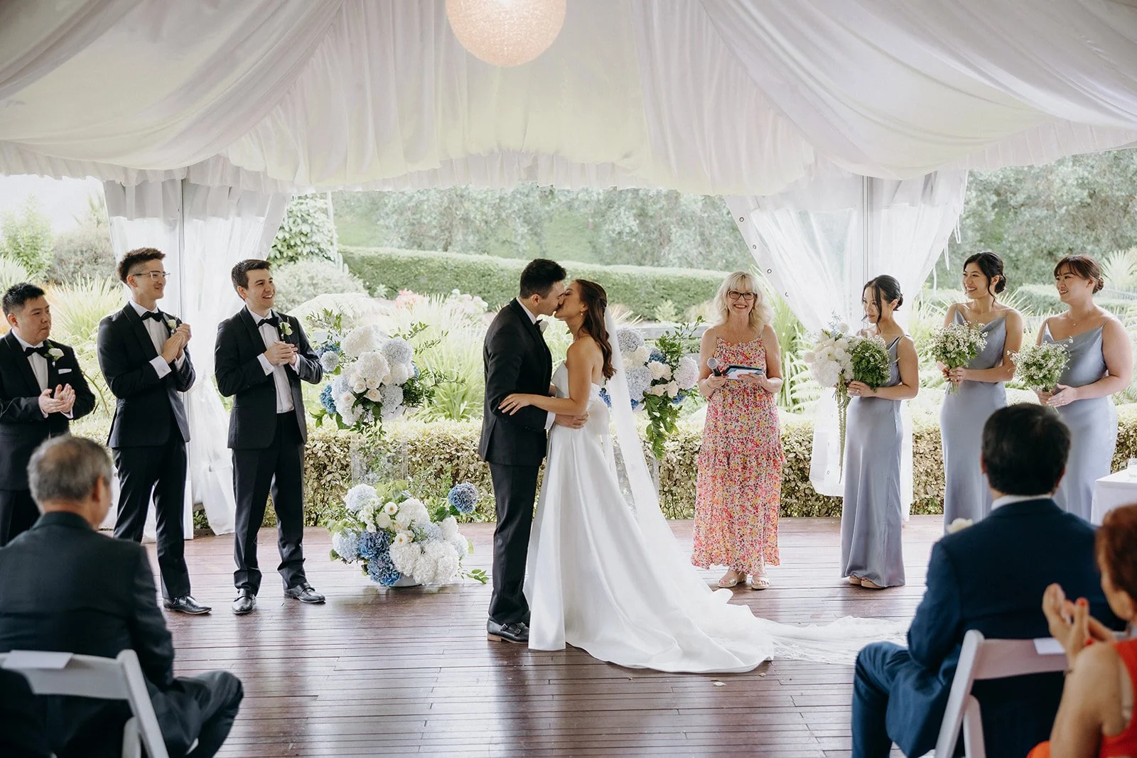 A wedding ceremony with a bride and groom kissing under the white canopy of the Marquee, surrounded by bridesmaids holding bouquets and groomsmen in suits, with guests seated watching outdoors.