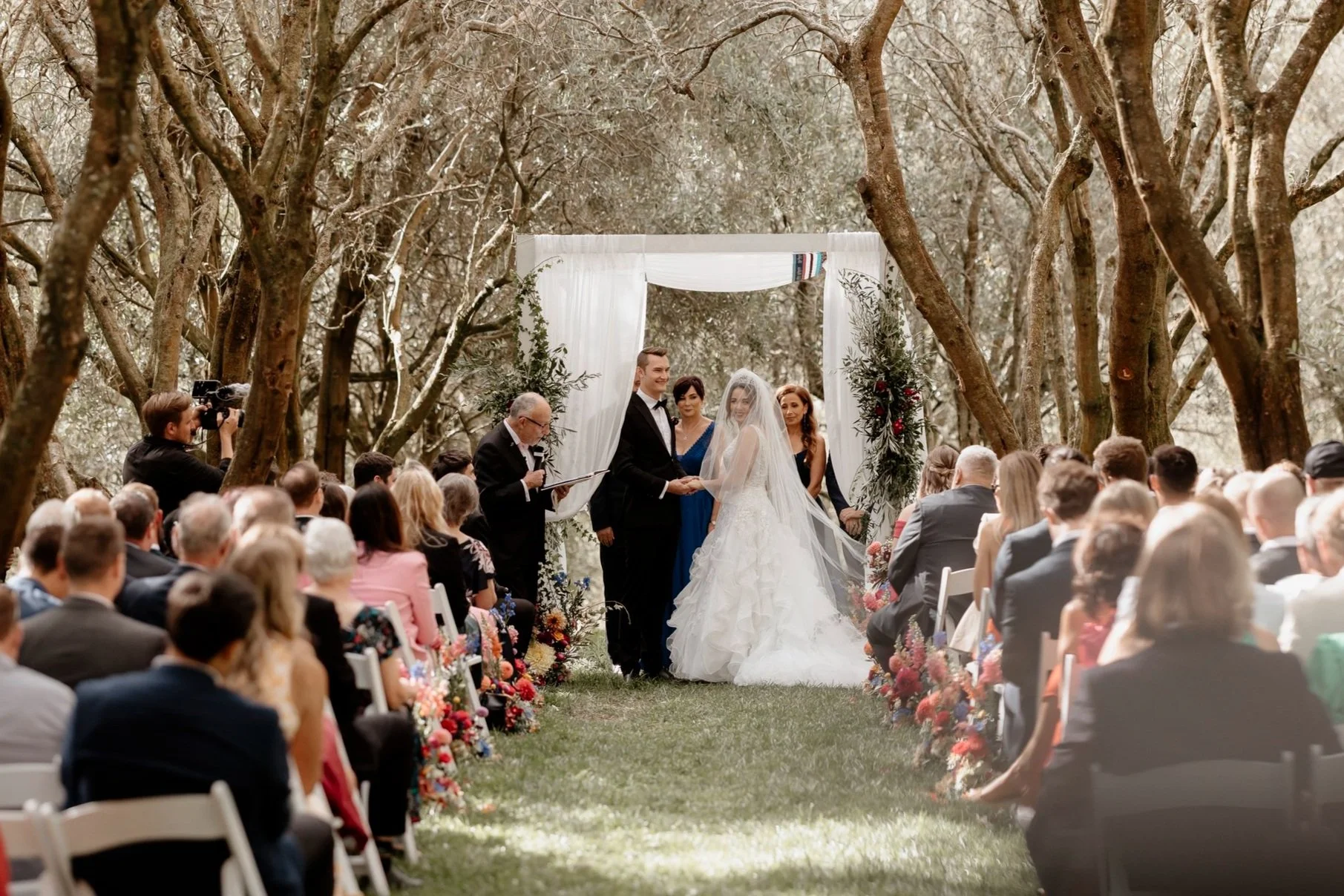 A wedding ceremony outdoors in Bracu's Olive Groves with guests seated facing the bride and groom under a white canopy decorated with greenery and flowers.