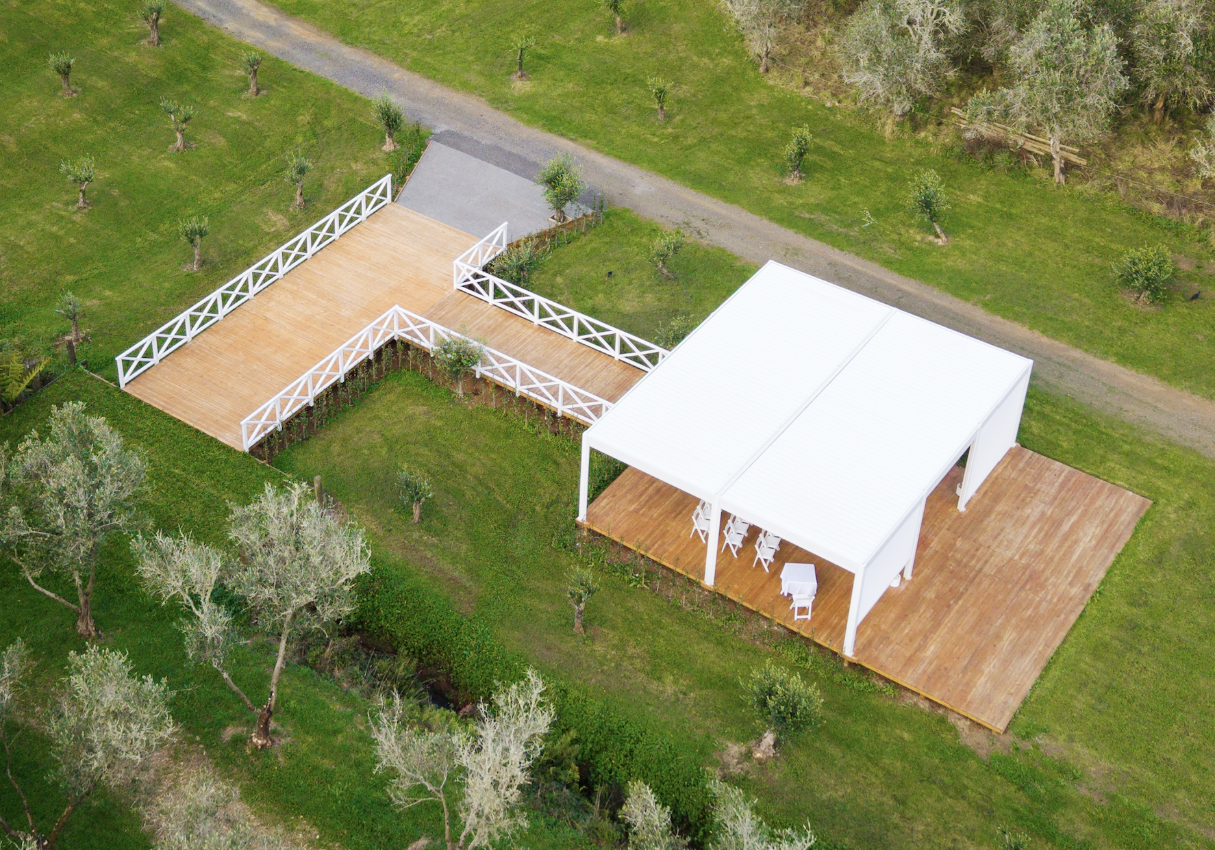 Aerial view of our newest wedding venue, the Velarium. Boasting a white louvred roof and a large wooden deck, with a white pergola, surrounded by green grass and trees.