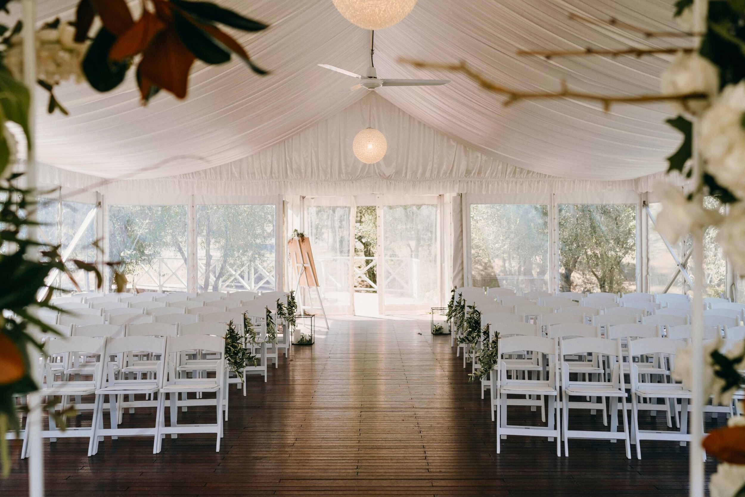 Empty wedding ceremony setup inside the Marquee, a white tent with white chairs decorated with greenery, a wooden floor, and a backdrop of trees.