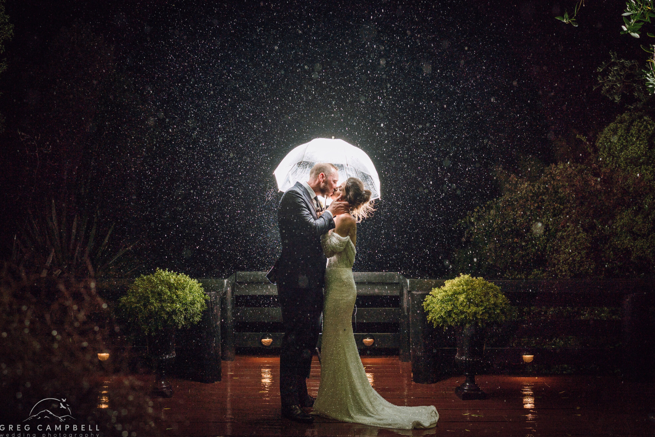 A bride and groom sharing a kiss under an umbrella during a rainstorm at night, illuminated by rain and possibly camera flash on a wooden deck with potted plants around them.