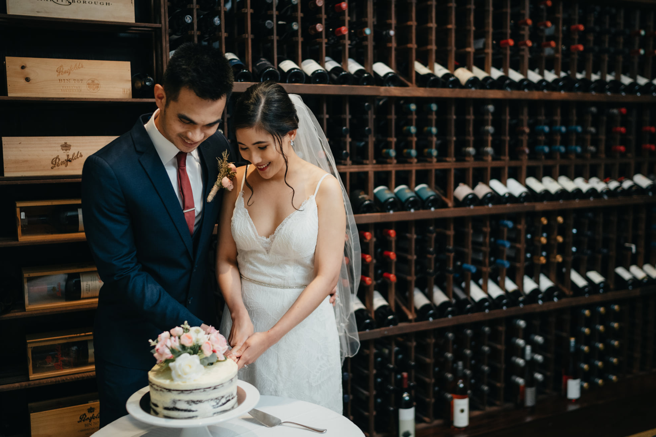 A bride and groom cutting a wedding cake together in The Wine Library, with wine bottles on wooden racks behind them.