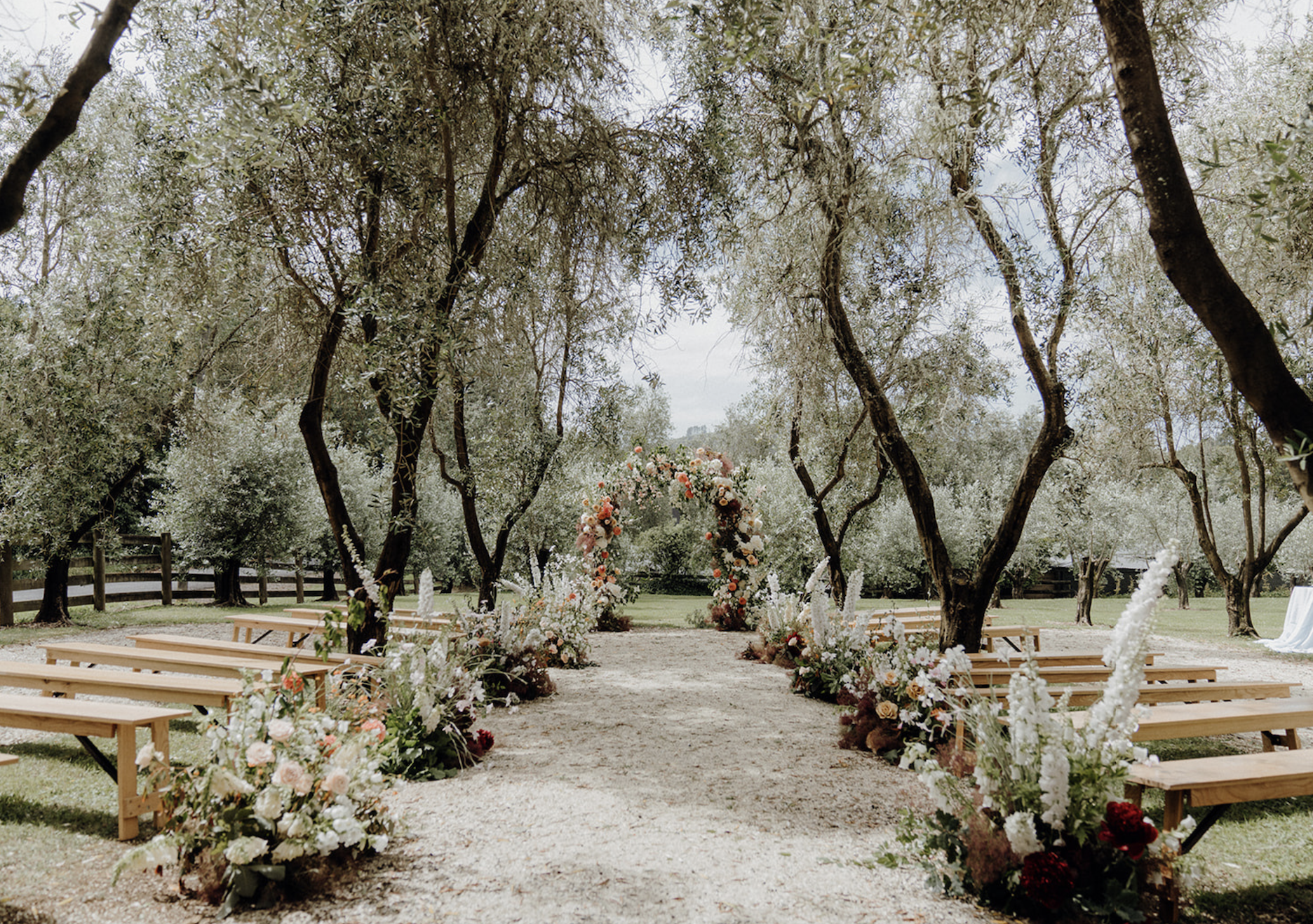 Outdoor wedding ceremony setup with wooden benches, floral arrangements, and arch amidst trees.