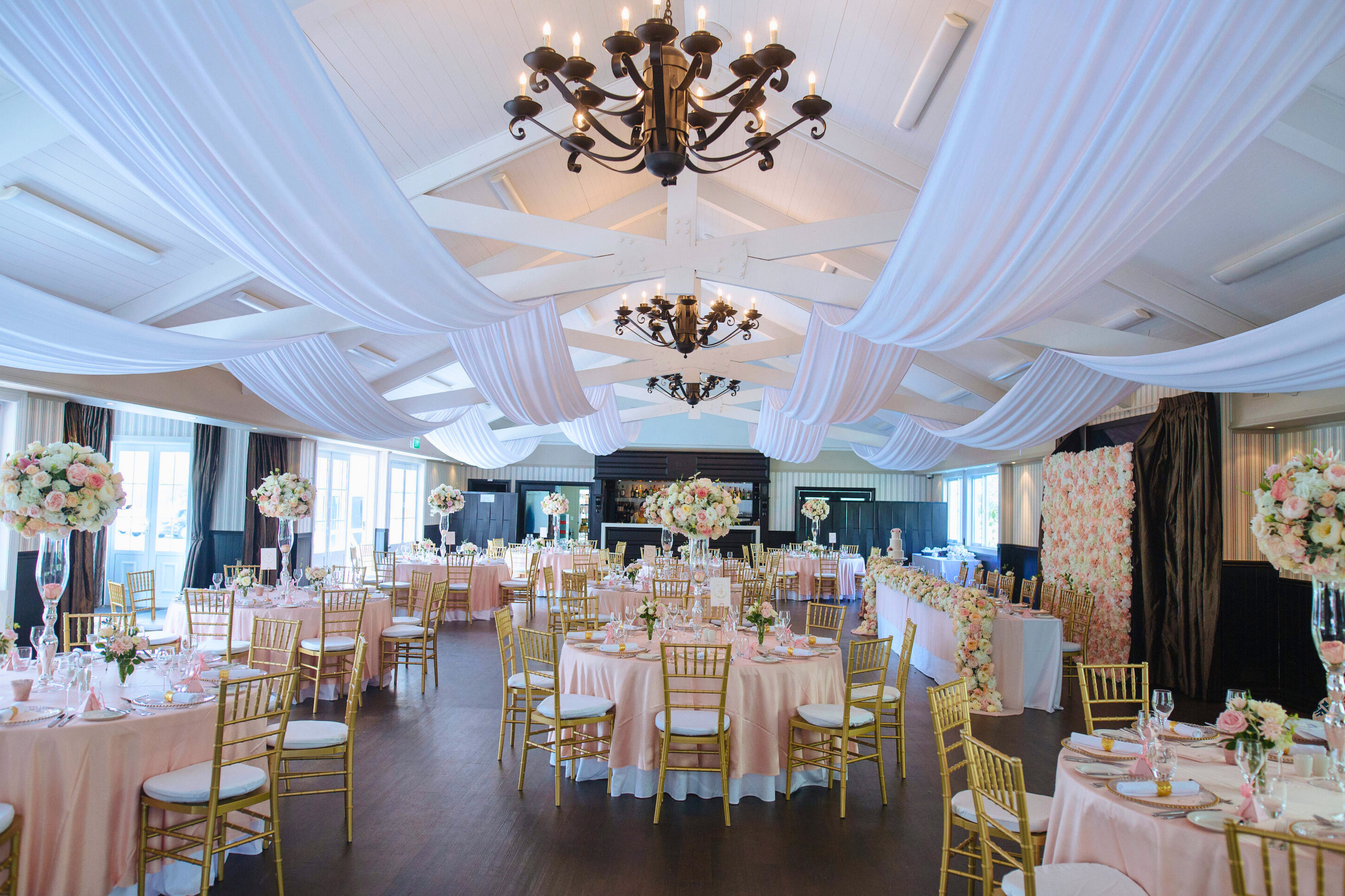 Elegant banquet hall (The Pavilion) decorated for an event with draped white fabric on the ceiling, black chandeliers, and round tables covered with pink tablecloths, floral centrepieces, and gold chairs.
