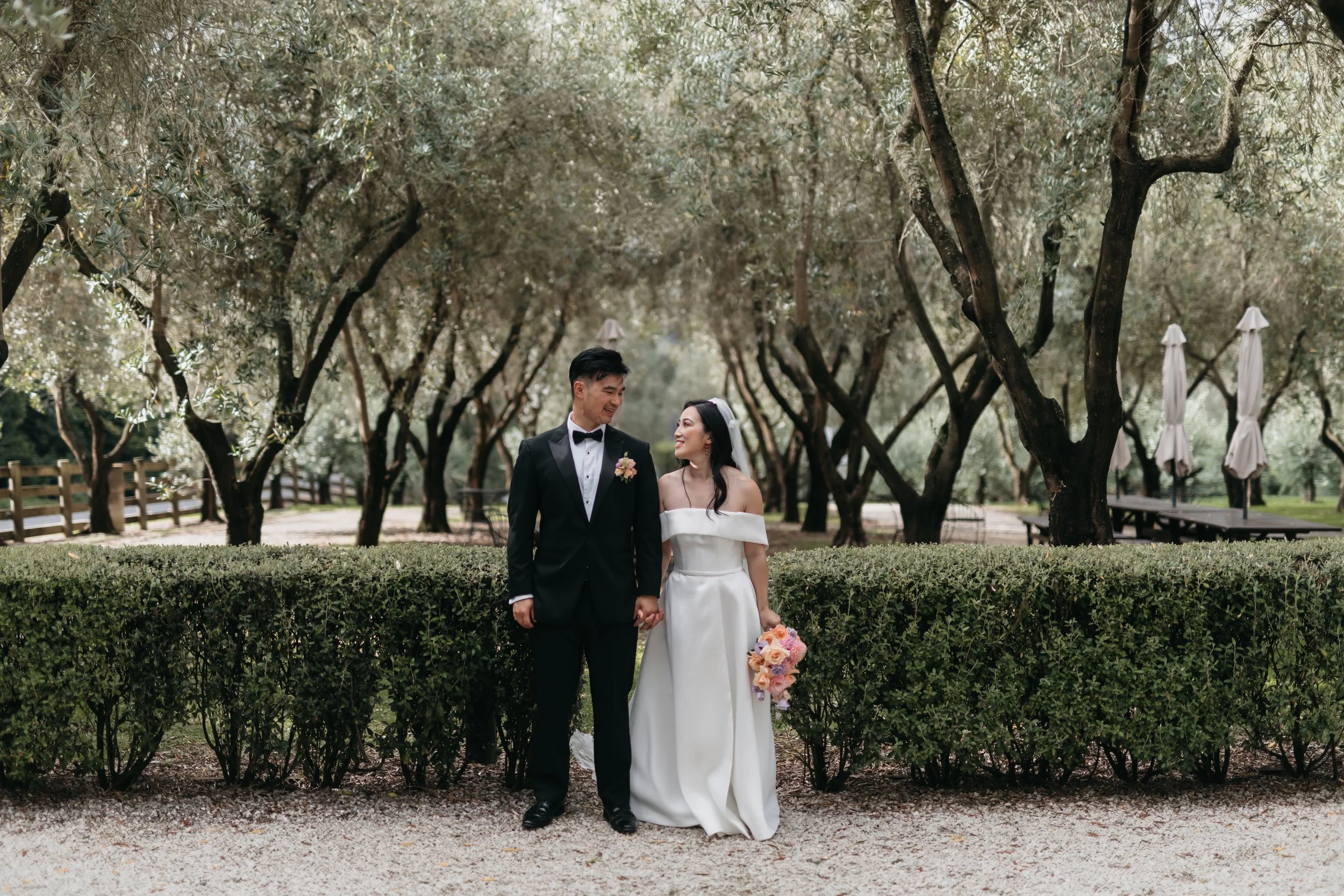 A bride and groom standing hand in hand outdoors among trees, smiling at each other during their wedding day. The bride wears a white off-shoulder gown and holds a bouquet of pink and white flowers, while the groom wears a black tuxedo with a bowtie and boutonniere.
