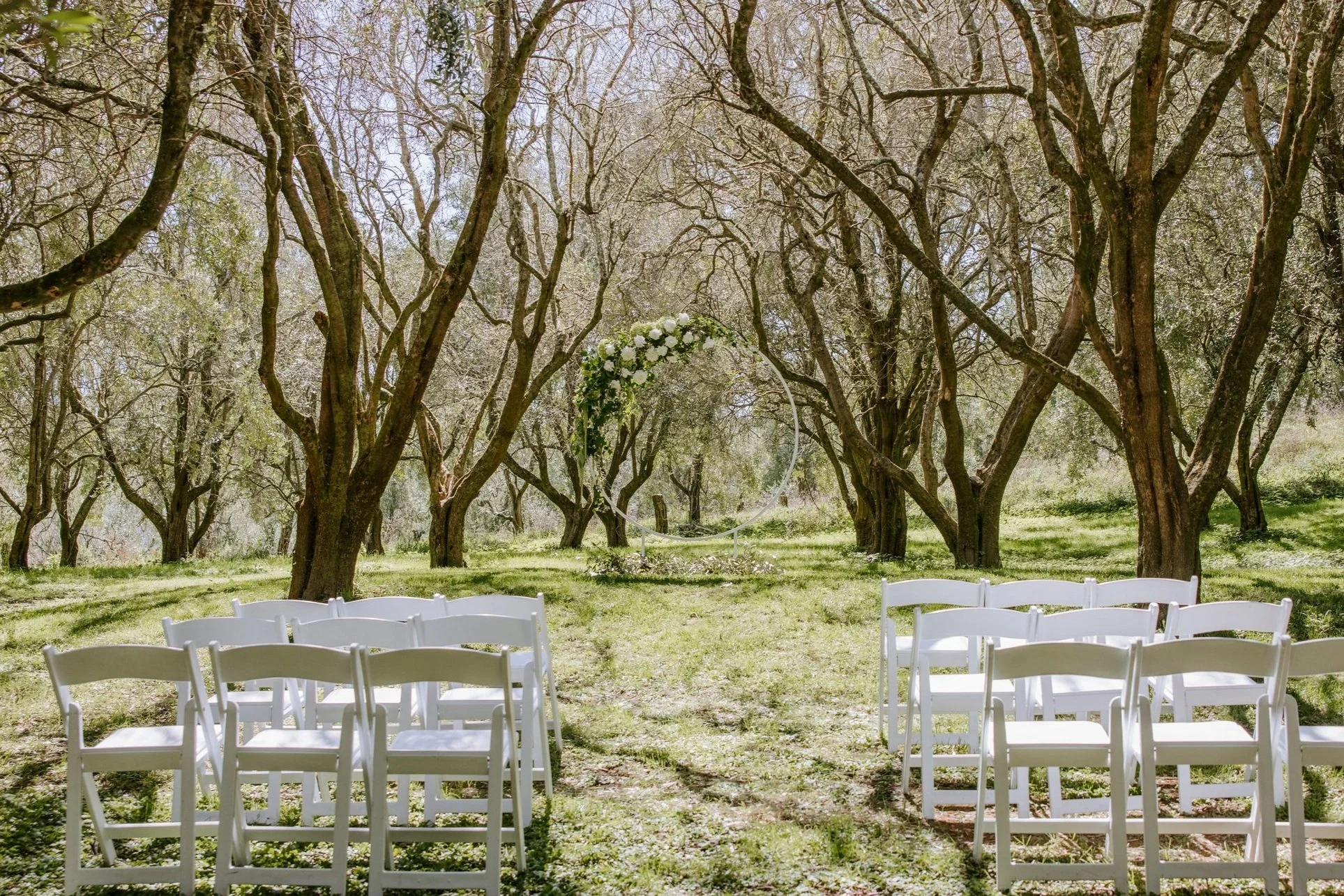 Outdoor wedding ceremony setup in a forest with white folding chairs arranged on grass, a circular floral arch, and tall trees with lush green leaves.
